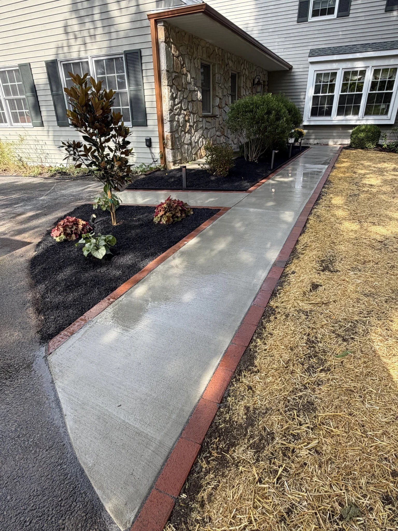 Residential concrete walkway with red brick edging leading to a home entrance, surrounded by landscaped garden beds with black mulch and ornamental plants.
