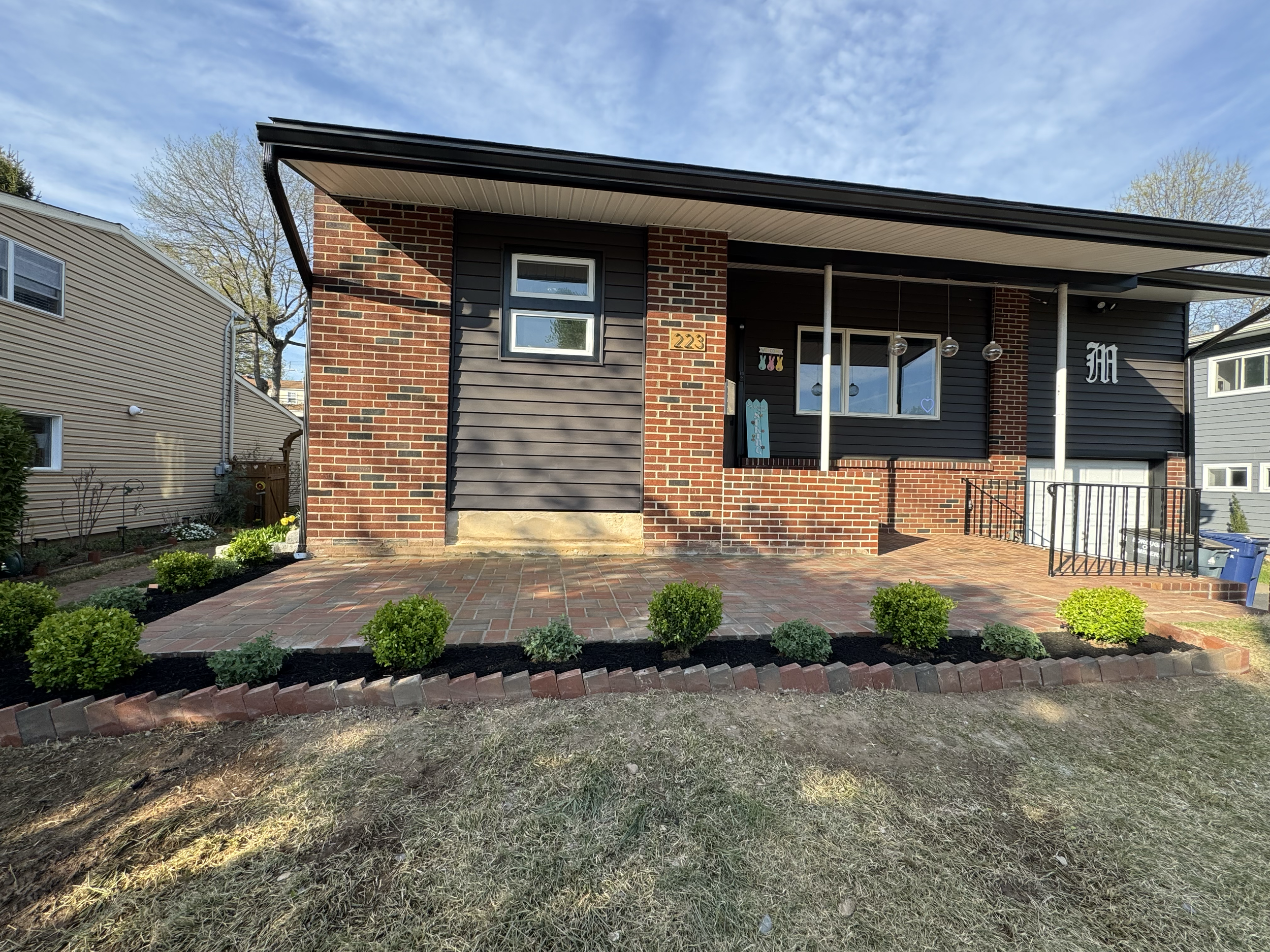 Front view of a modern house with brick and dark siding exterior, a small front porch with hanging lights, a garden with bushes, and a paved walkway, under a blue sky with scattered clouds.