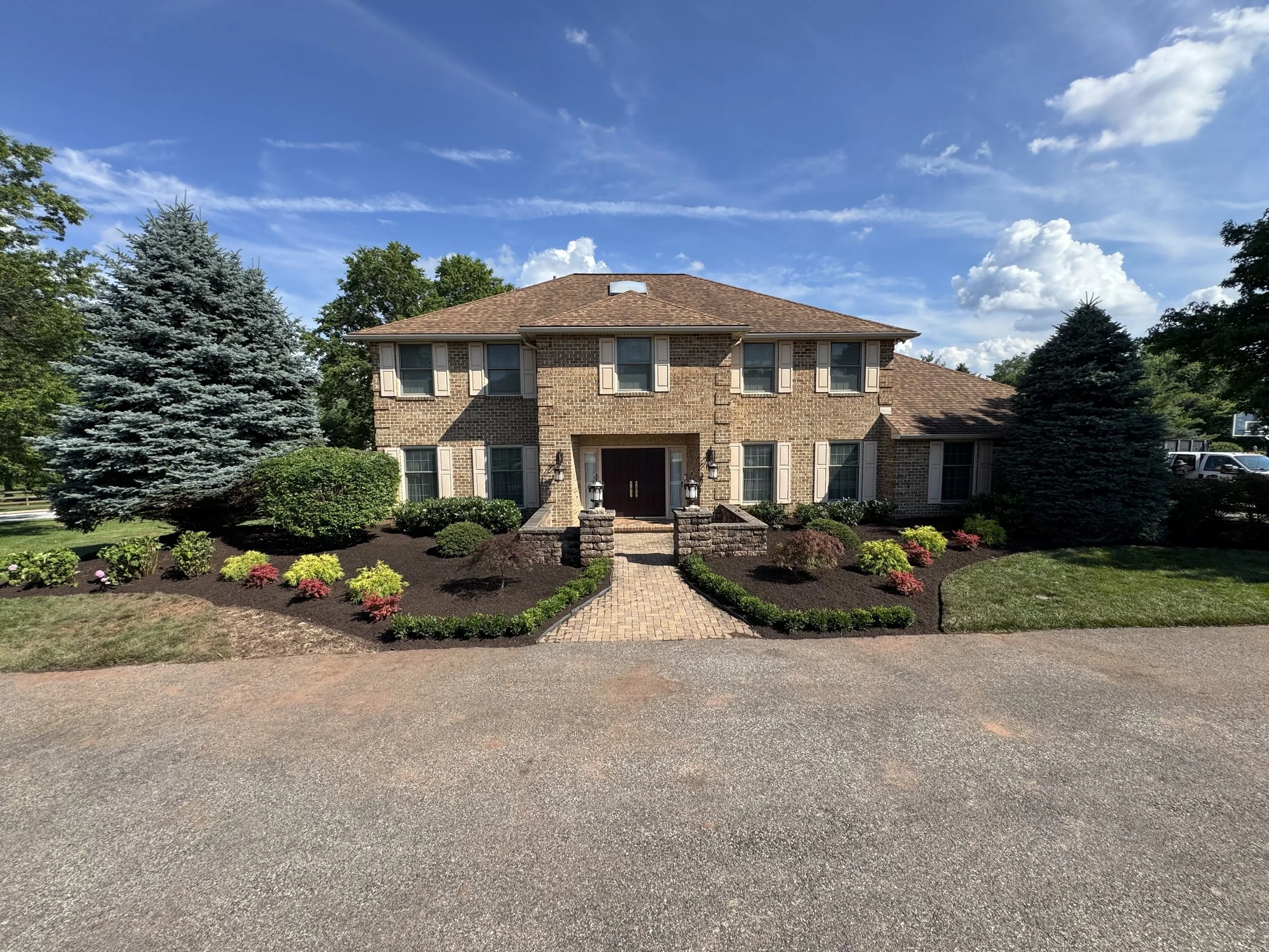 Front yard landscaping with fresh black mulch, colorful ornamental shrubs, mature evergreen trees, and a paver walkway leading to a brick home.