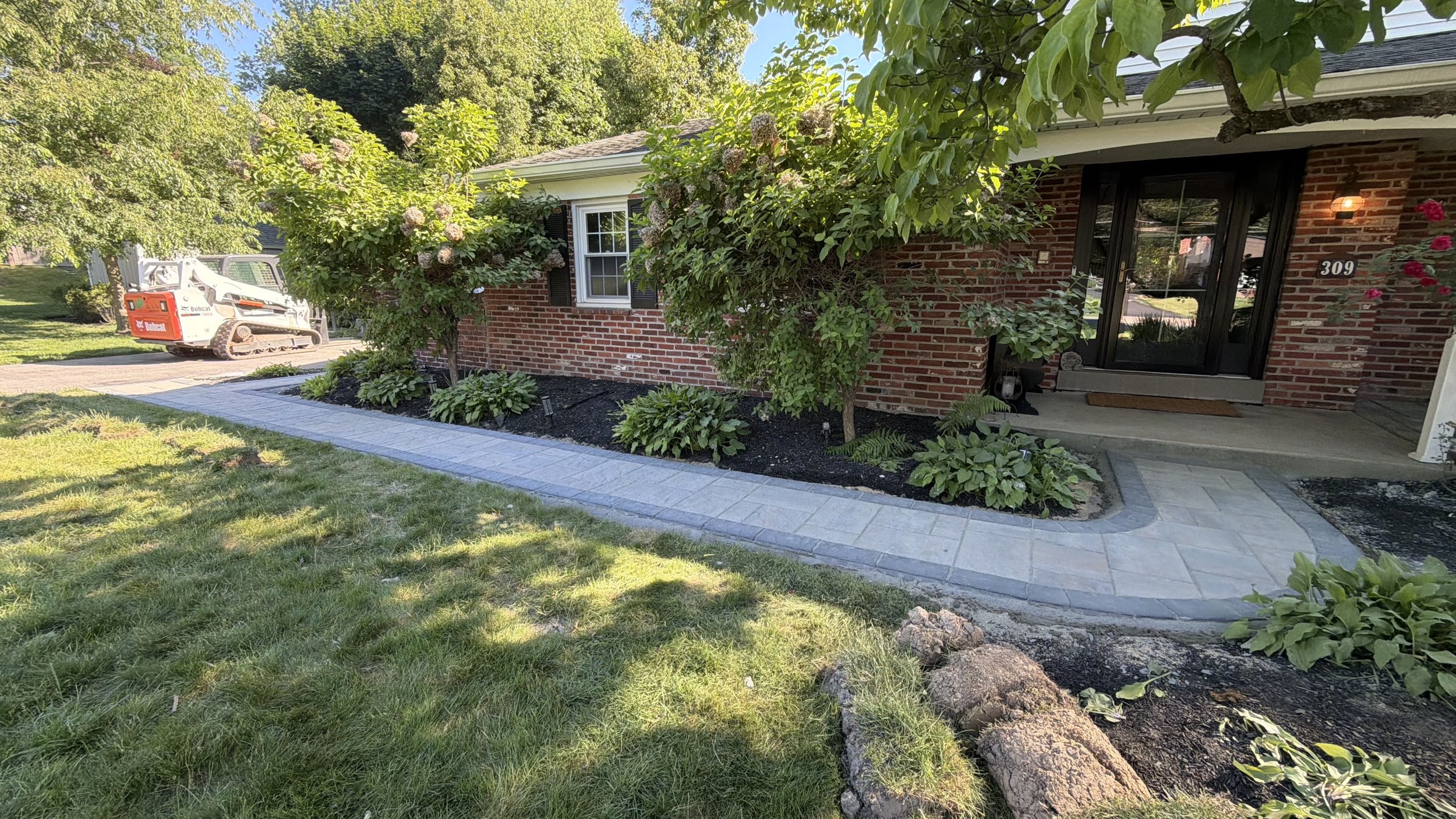 Custom paver walkway along a brick home, featuring a clean stone border, established shrubs, and landscaped planting beds that create an inviting entrance and complement the property's exterior.
