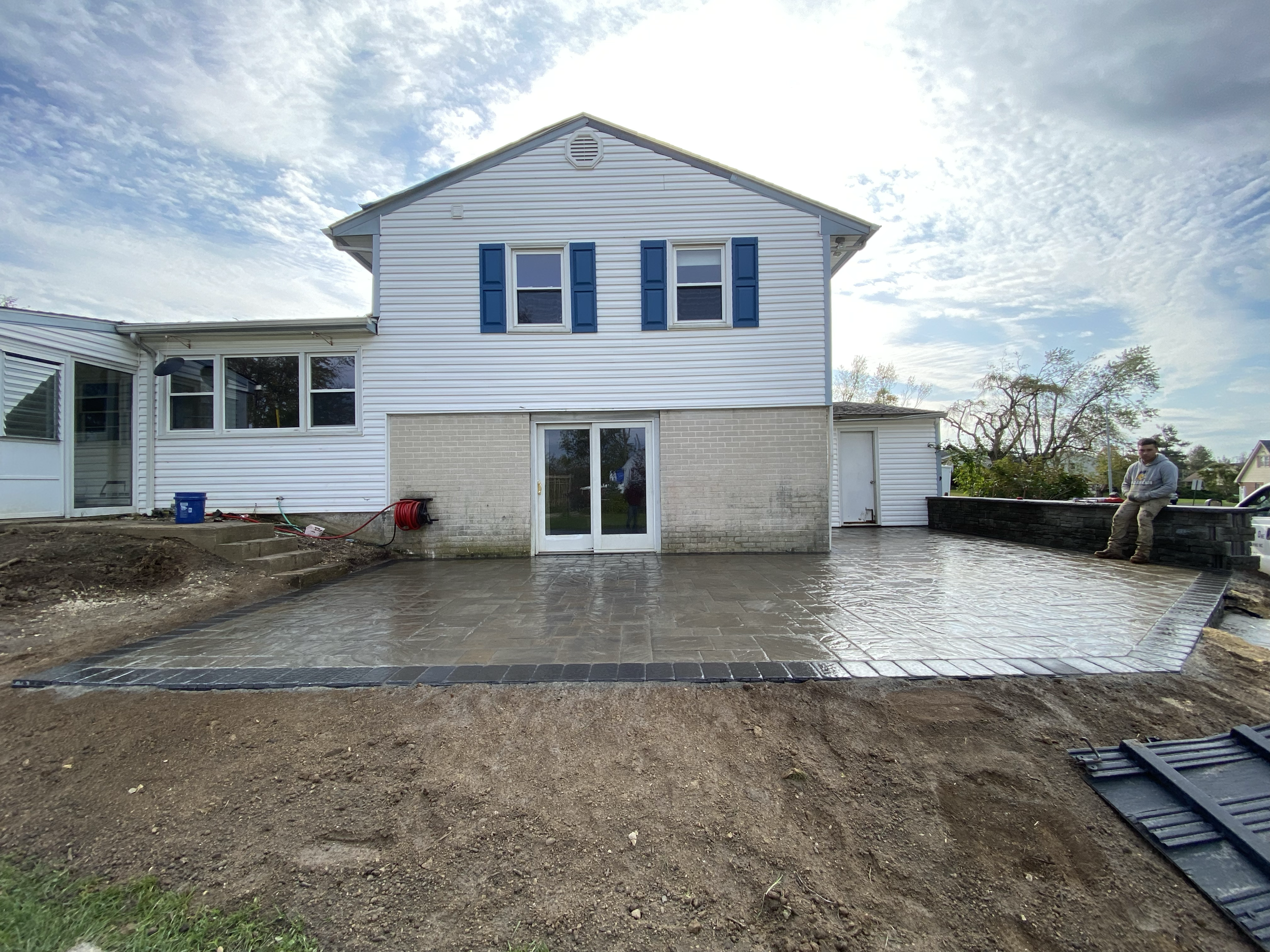 Backyard view of a house with white siding, blue shutters, a sliding door, and a brick foundation, with a newly paved patio area and some construction tools and dirt in the foreground.