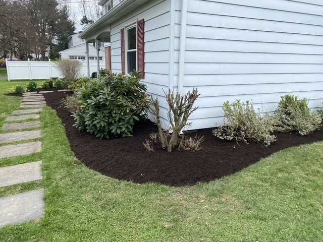 Residential front yard landscaping featuring fresh brown mulch, ornamental shrubs, decorative plants, and neatly maintained garden beds along the front of a stone house.