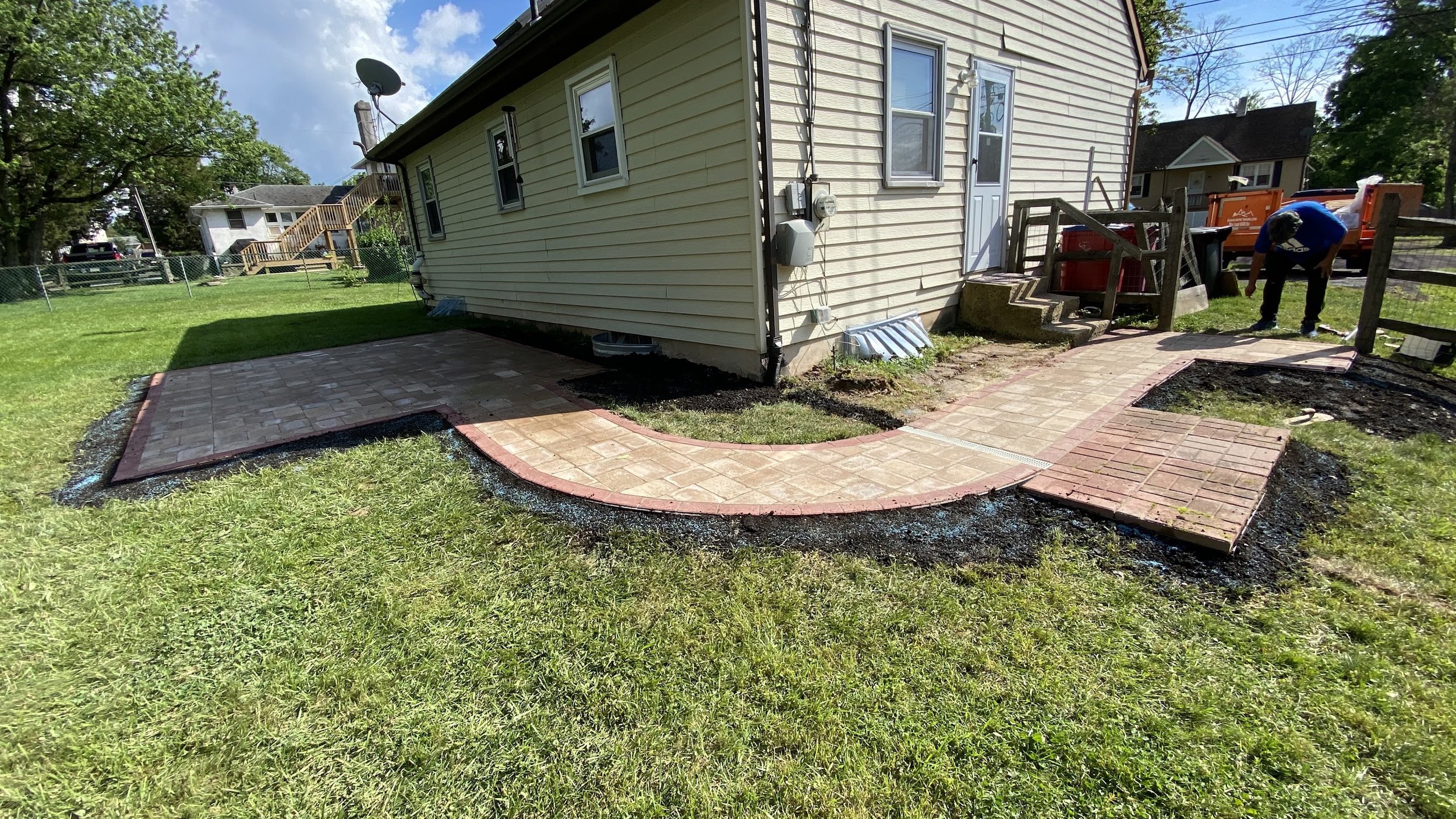 Backyard with new brick patio and brick pathway around the house, yellow siding, stairs to the deck, and a worker laying bricks.
