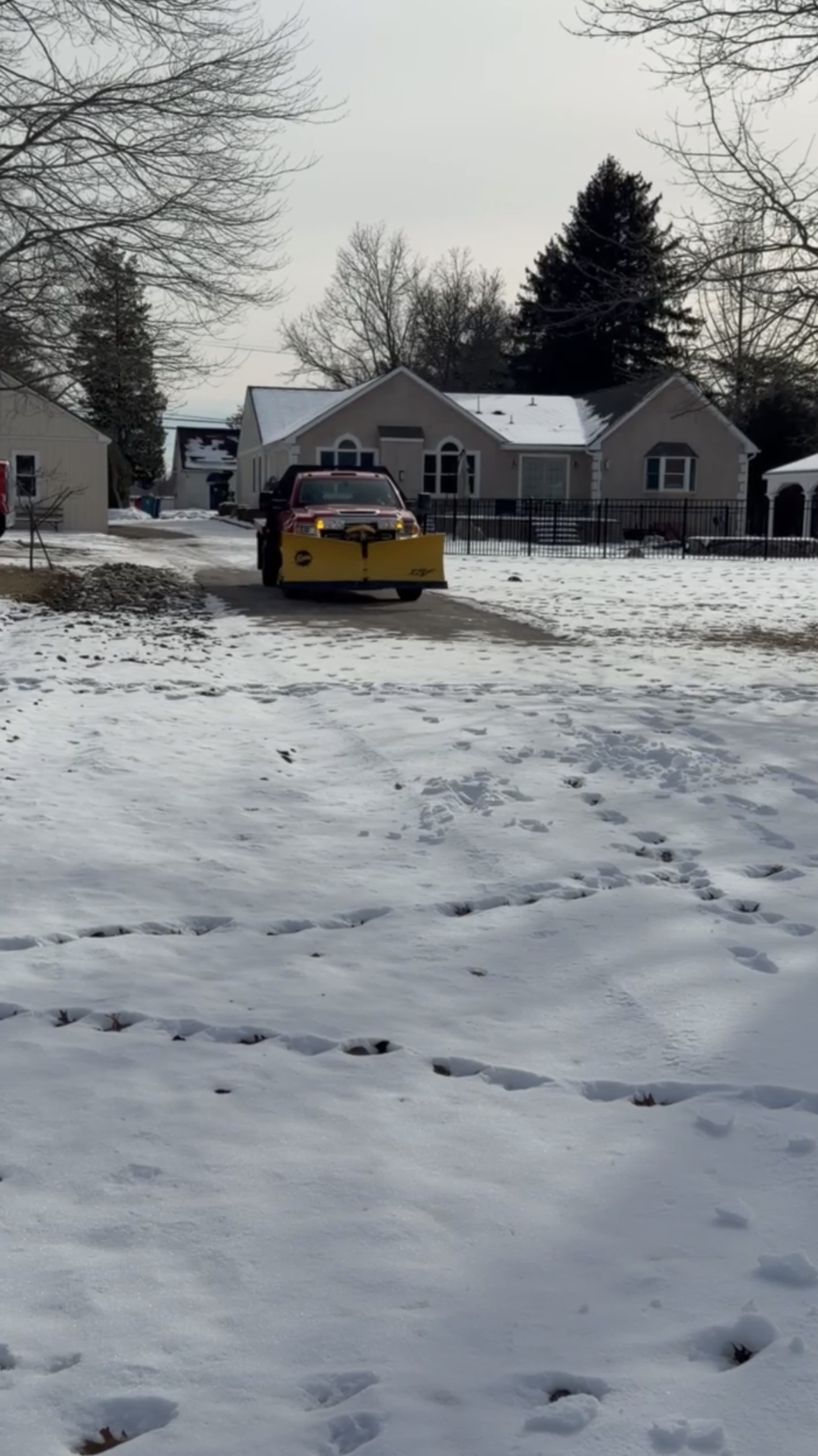 A snow-covered suburban yard with tracks and footprints in the snow. A truck equipped with a snowplow blade is on the driveway. Houses and trees are visible in the background, with one house having a fence around the yard.