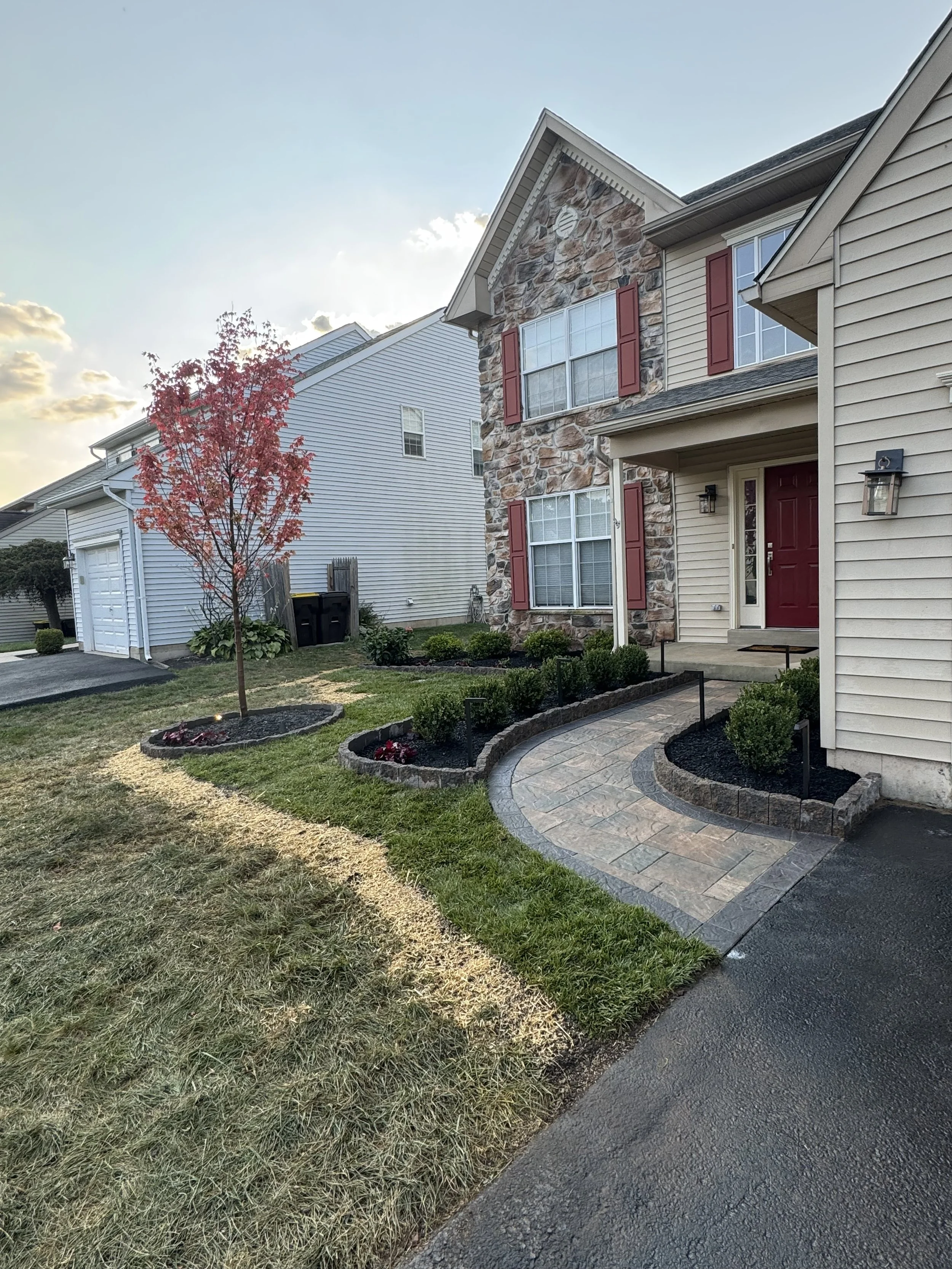 A suburban house with stone and siding exterior, red shutters, red front door, manicured lawn with a tree and flower bed, and a paved walkway leading to the entrance.