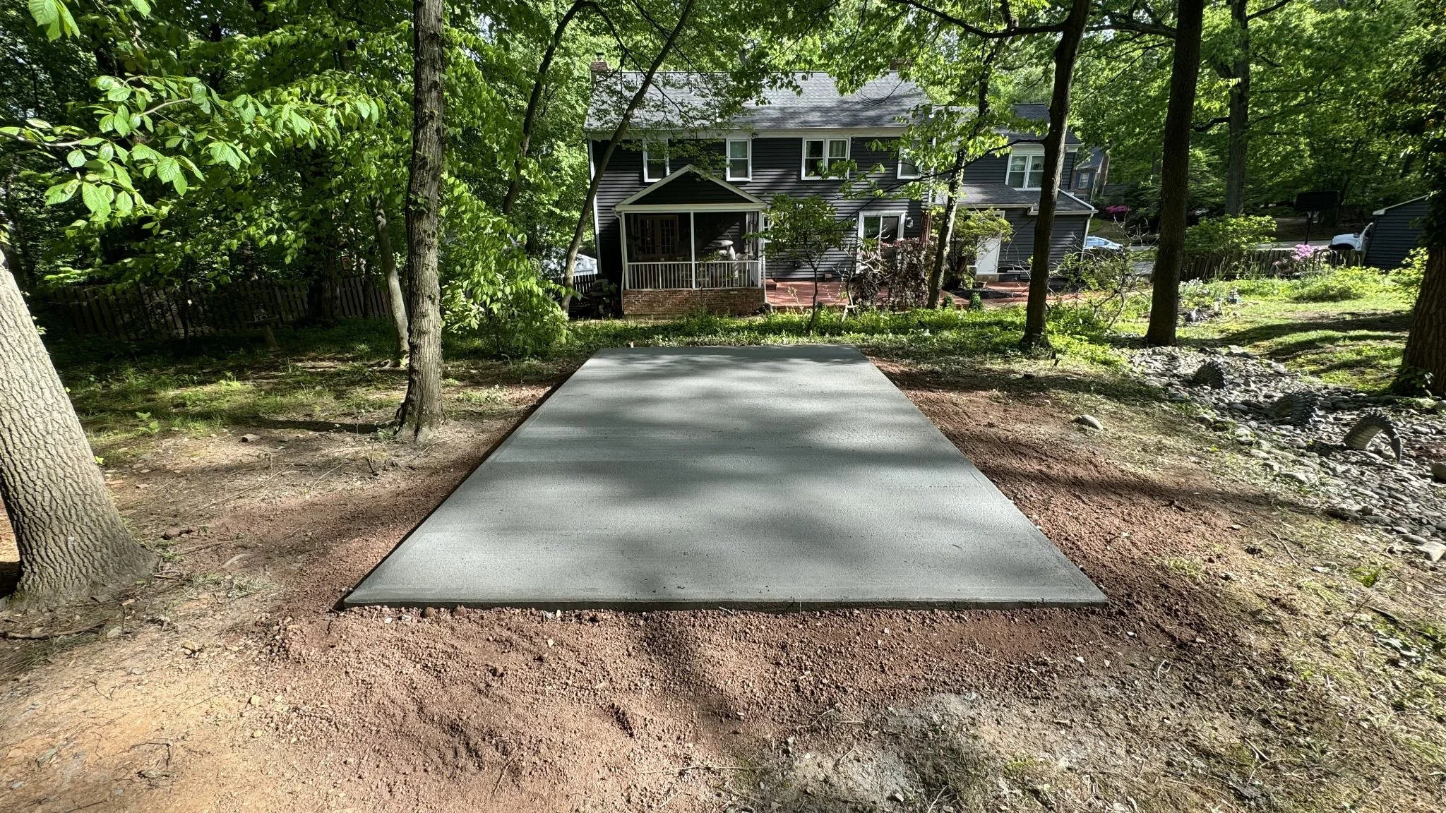 Newly installed rectangular concrete pad with a smooth finished surface in a residential backyard surrounded by mature trees.