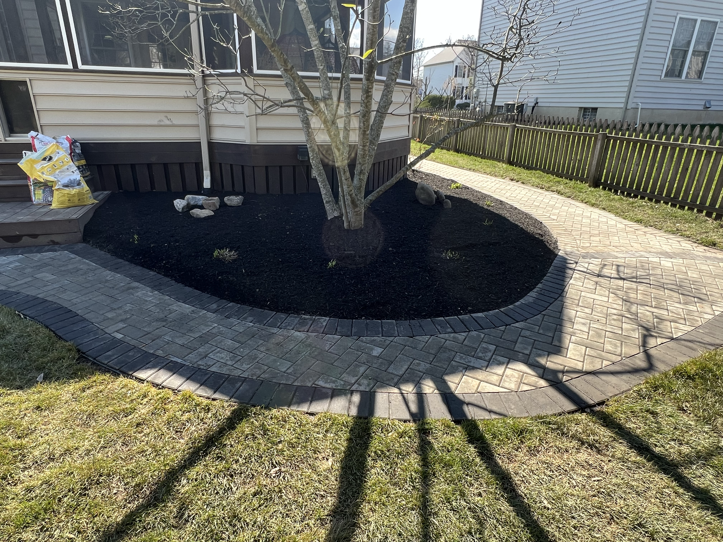 A backyard garden with a small tree planted in a mulched bed, surrounded by a stone paver patio. There are gardening supplies on a small wooden deck, and a shadow cast by the tree falls across the grass and patio. In the background, there is a beige 