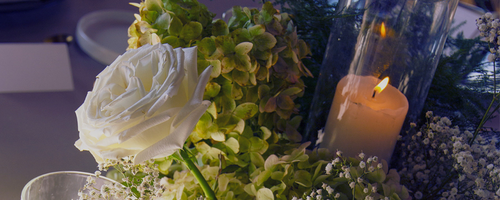 A floral arrangement featuring white roses and green hydrangeas with lit candles in glass holders in the background.