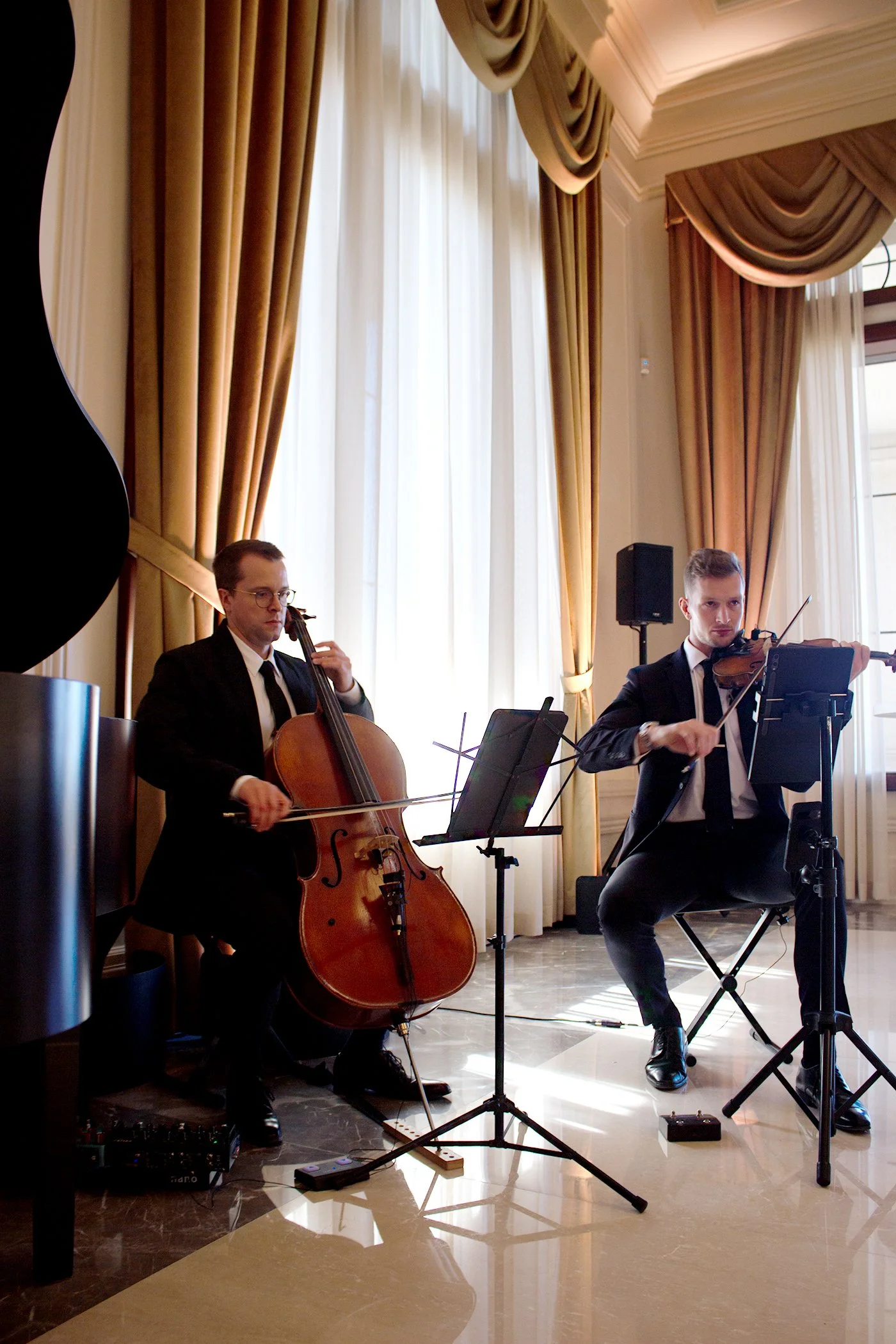 Two musicians in formal attire performing in a room with large golden curtains and natural light. One is playing a cello, the other a violin, with music stands and equipment around them.