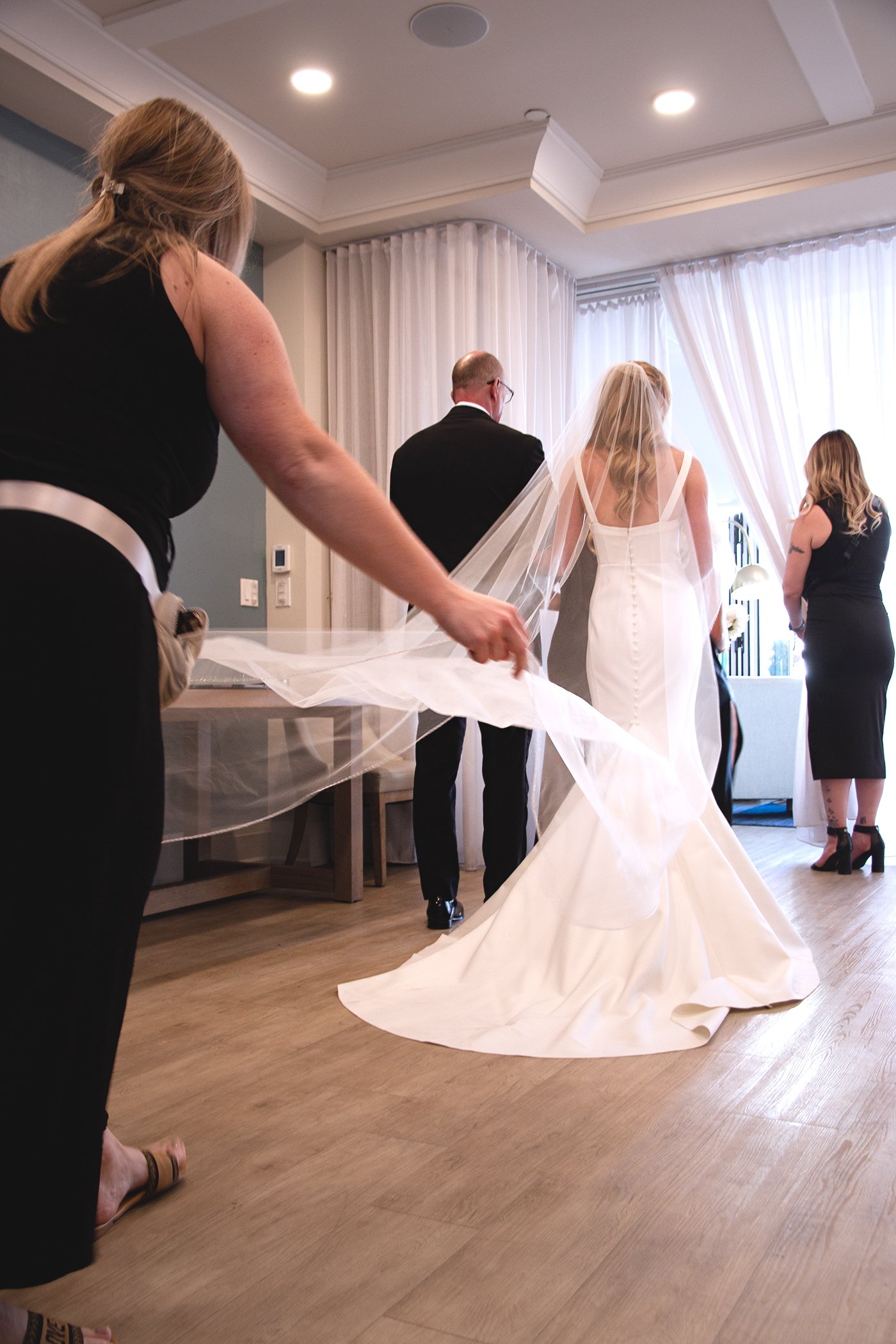 Bride in a white wedding gown with a long train and veil standing in a room with curtains, surrounded by women preparing for the wedding, one of whom is adjusting the train.