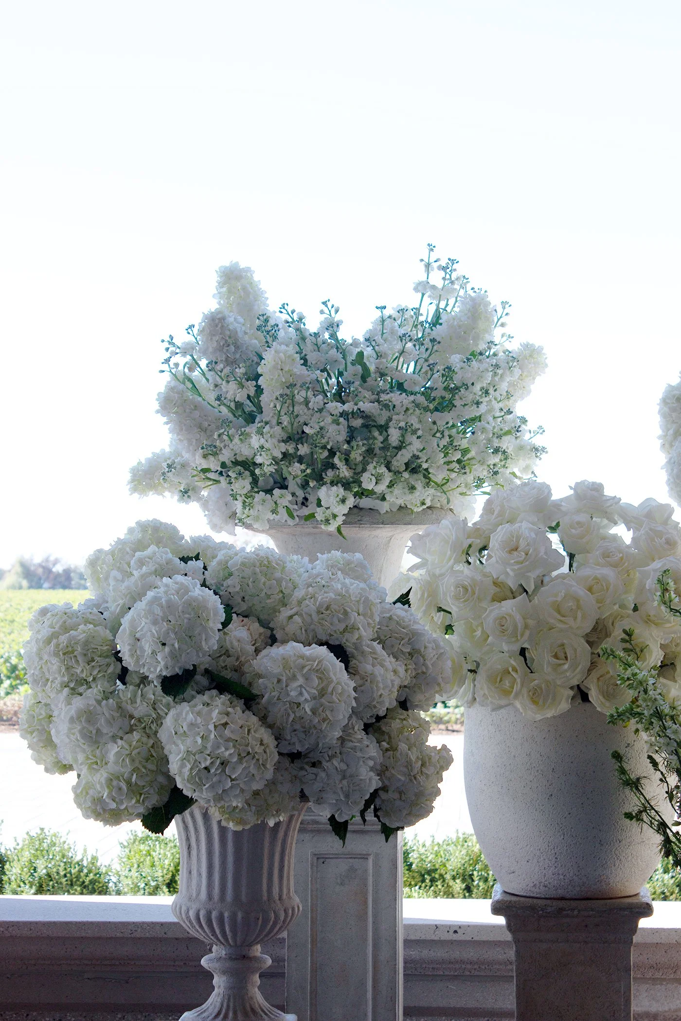 Close-up of two large white floral arrangements in decorative vases, positioned on a ledge outdoors with greenery and trees in the background.