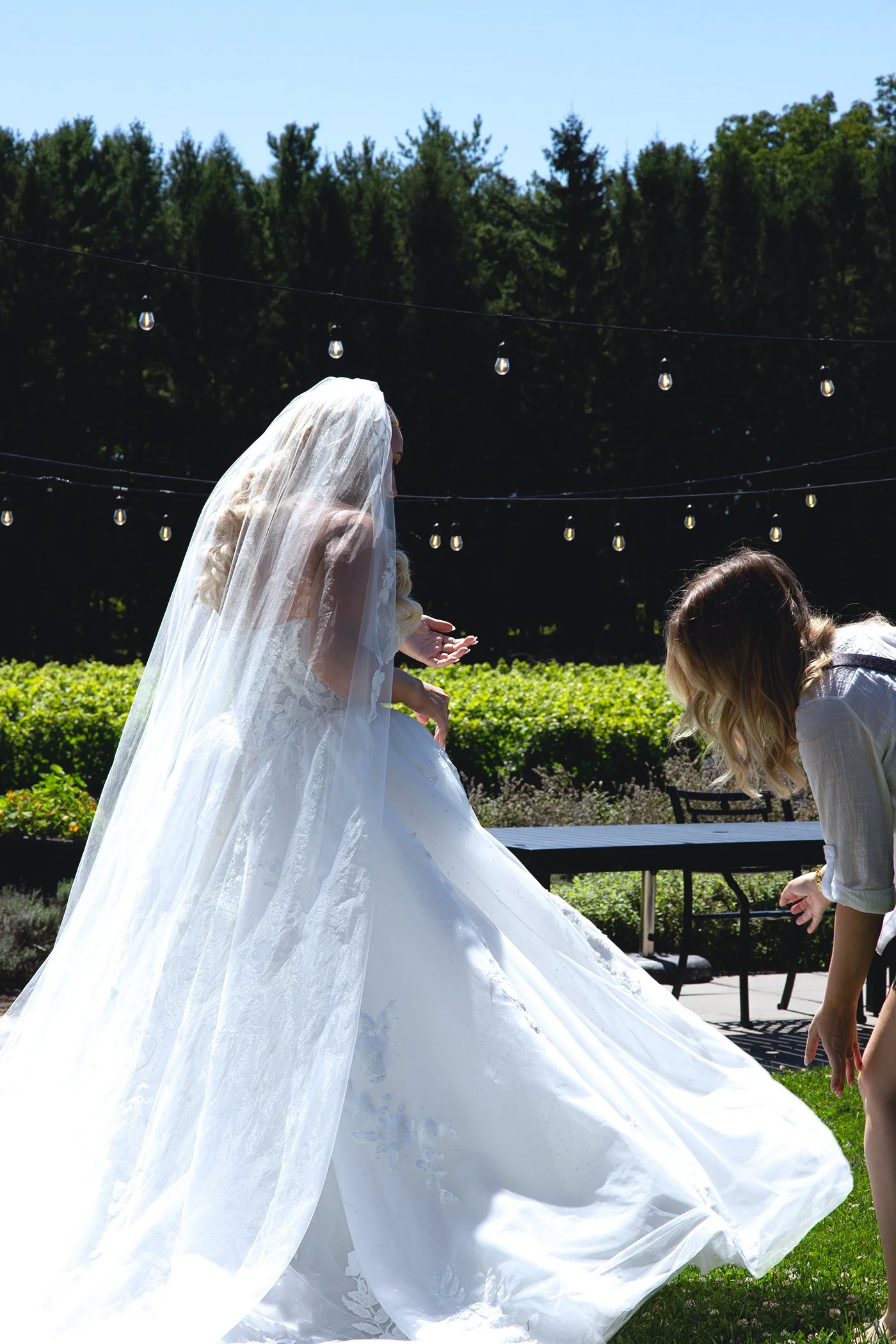 A bride in a white wedding dress and veil standing outdoors under string lights with greenery and trees in the background, assisted by a woman bending down.