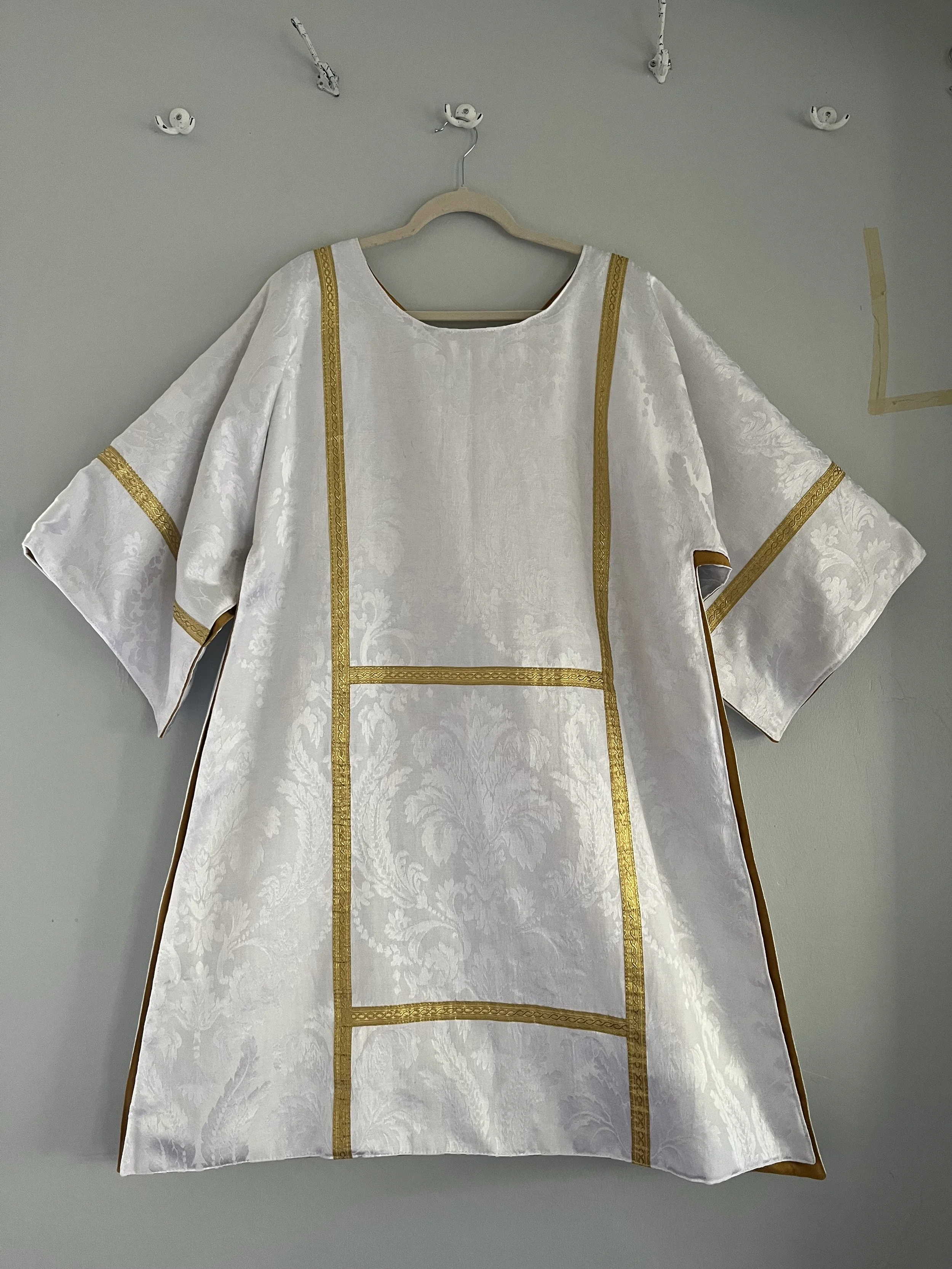 A white liturgical vestment with gold trim hangs on a wooden hanger against a plain light gray wall.