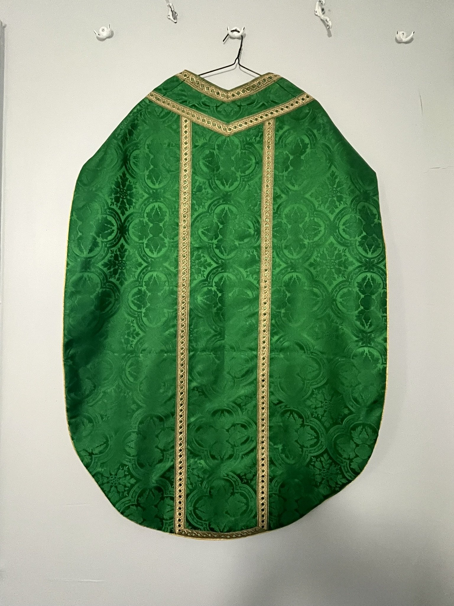 A green religious vestment with gold trim hanging on a wire hanger against a plain white wall.