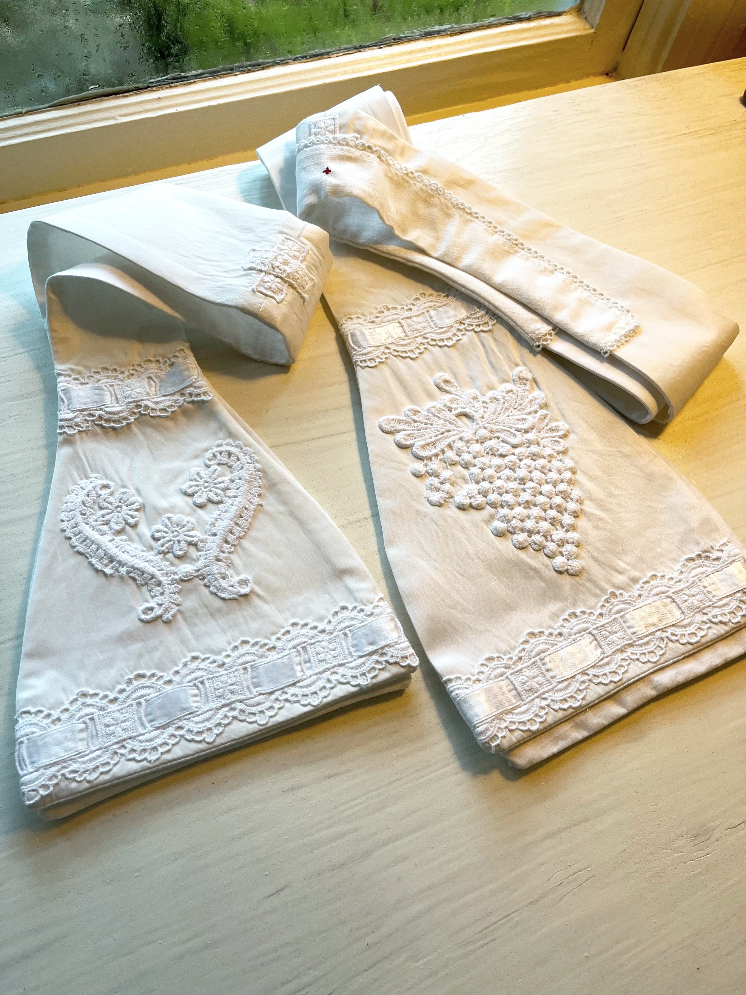 Folded white embroidered tablecloths or cloths with lace and embroidery, lying on a light wooden surface near a window