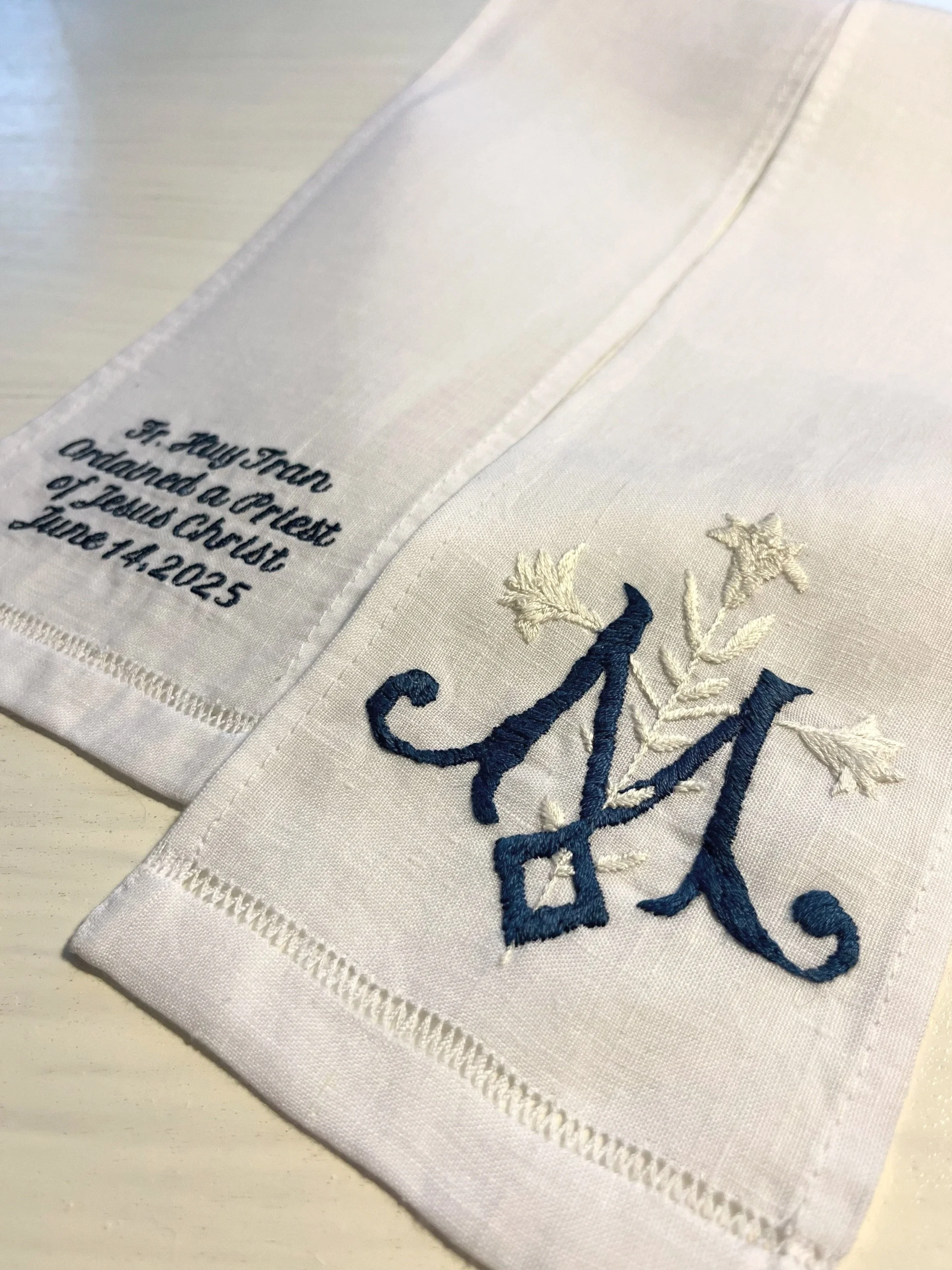 Close-up of a white cloth handkerchief with embroidered blue text and decorative embroidery, including the words 'St. Mary Talor' and an engraved date of August 14, 2025.