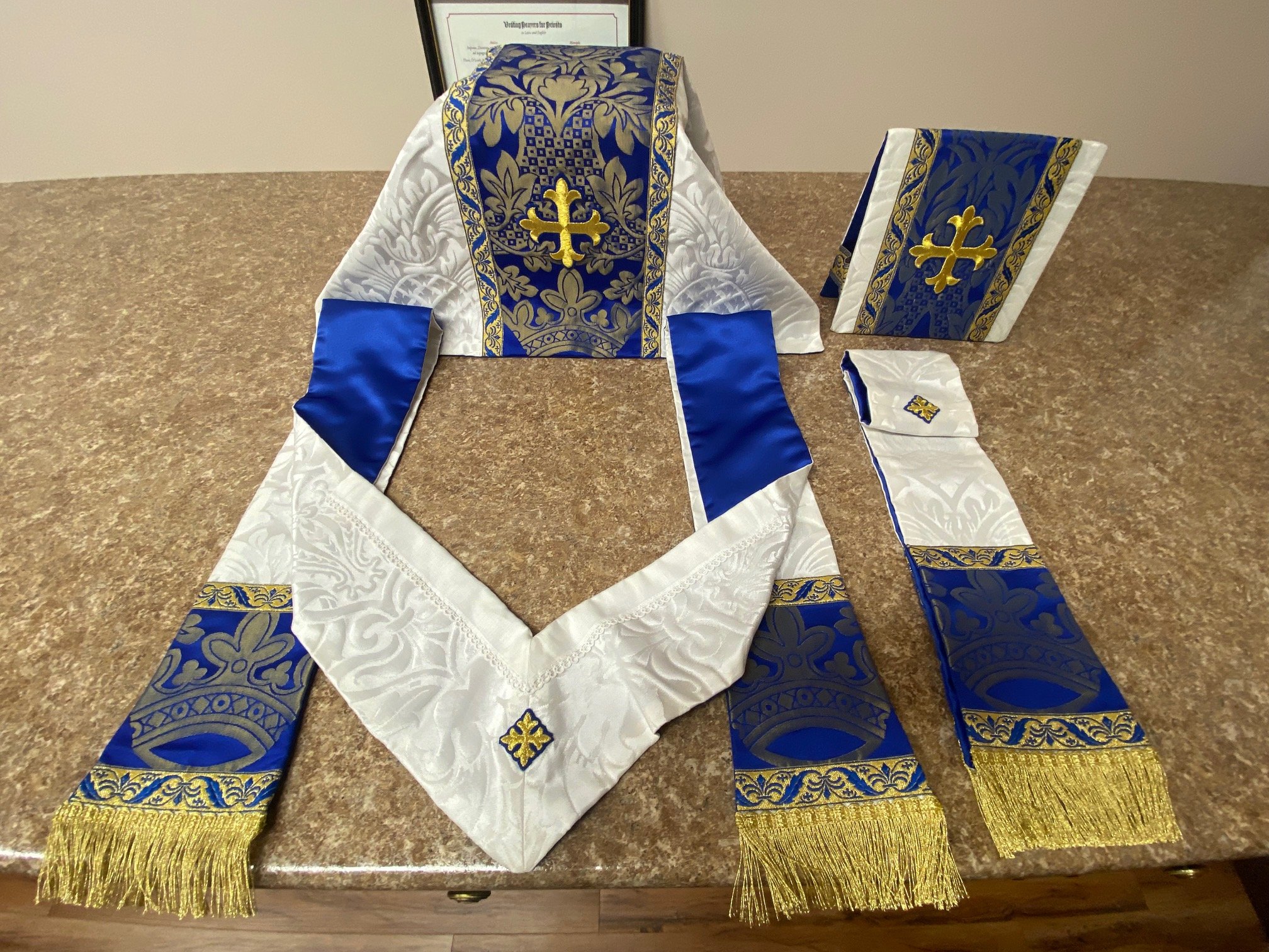 A collection of religious vestments and accessories, including a white and blue embroidered altar cloth, a matching stole or sash, a book cover or binder, and a framed certificate or document in the background, all with gold accents and fringe, displayed on a brown marble countertop.