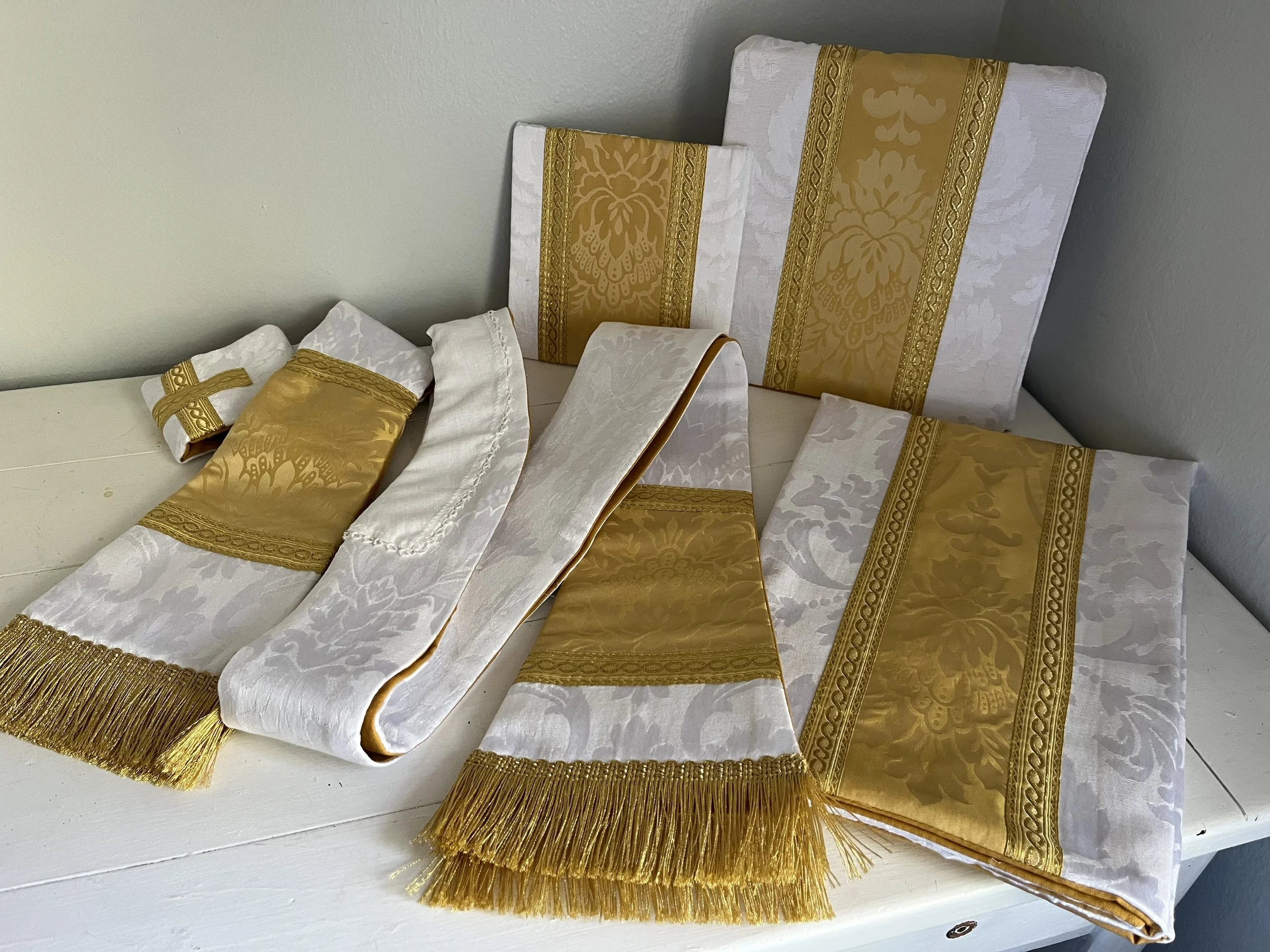 Set of white and gold embroidered and fringed cloths or table linens arranged on a white surface against a gray wall.
