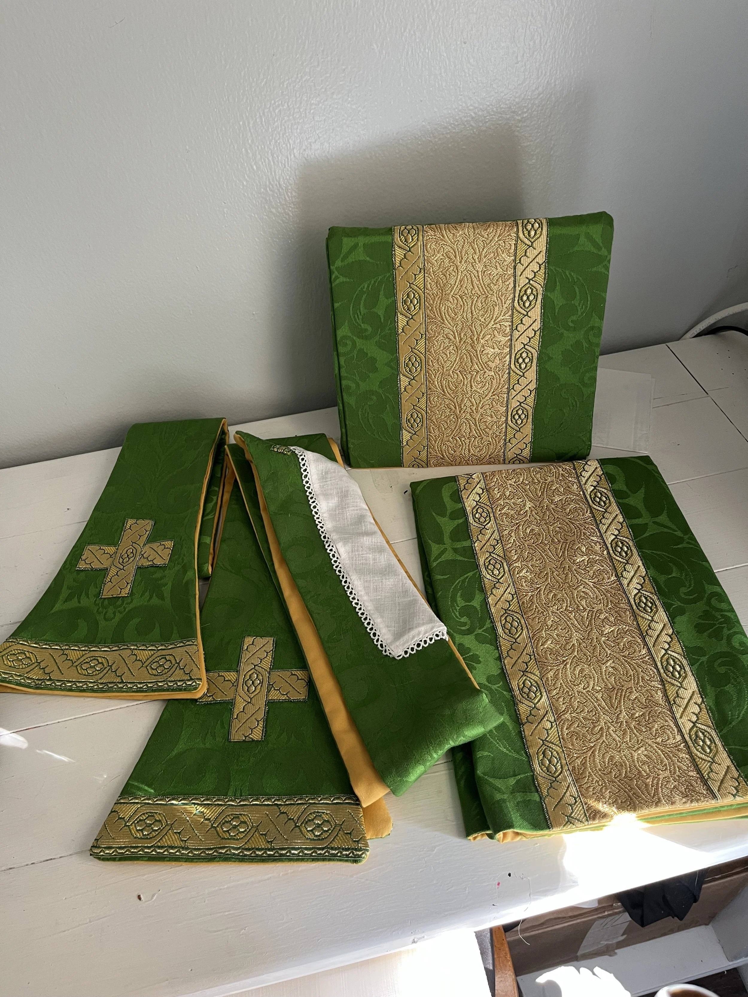 Green and gold fabric altar cloths with intricate patterns, some with gold crosses, laid on a white surface.