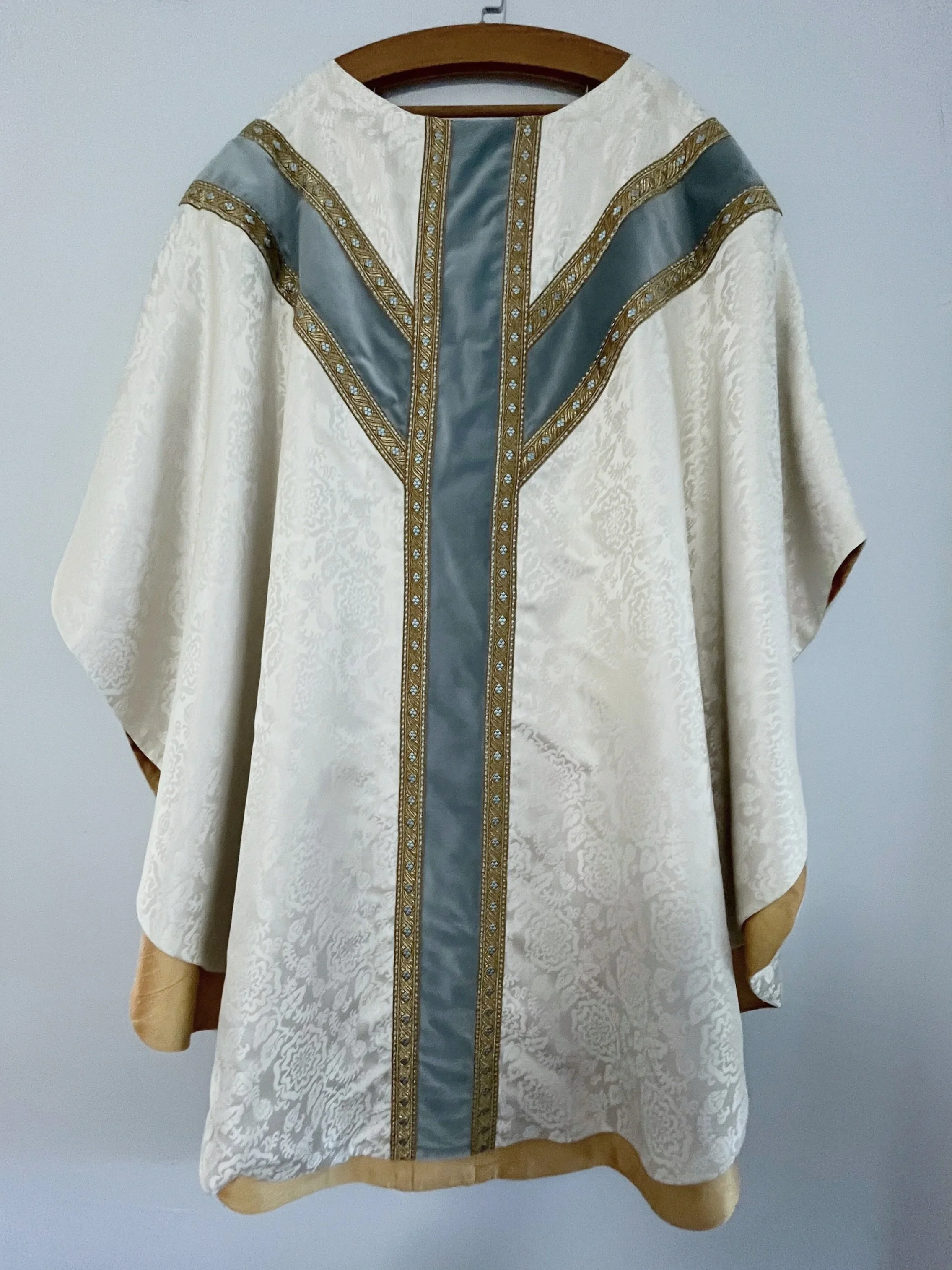A cream-colored ceremonial robe with gold and gray embroidered trim hanging on a wooden hanger against a gray wall.