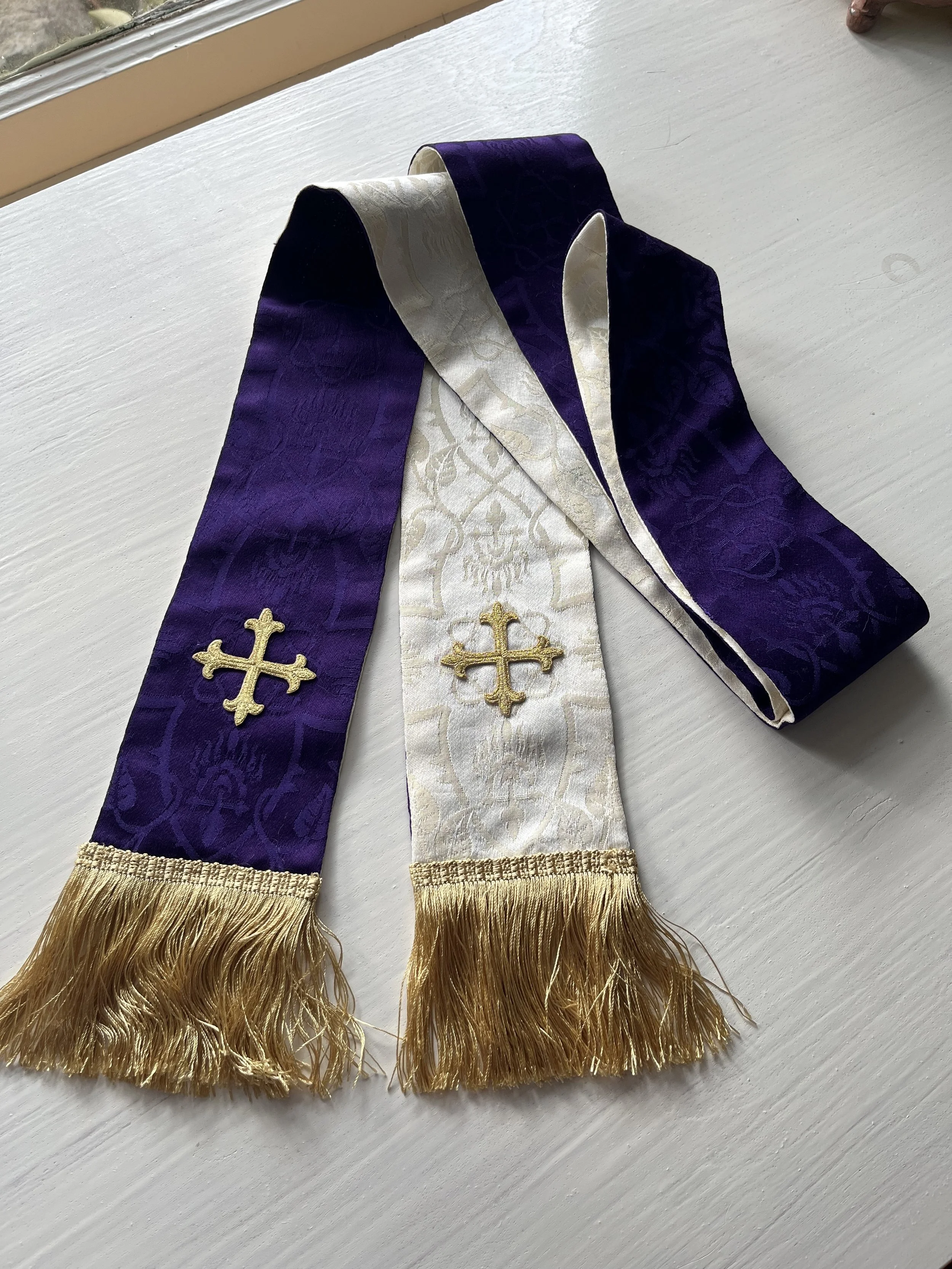 Three religious sashes with gold fringe and embroidered crosses, one white and two purple, laid on a white surface.
