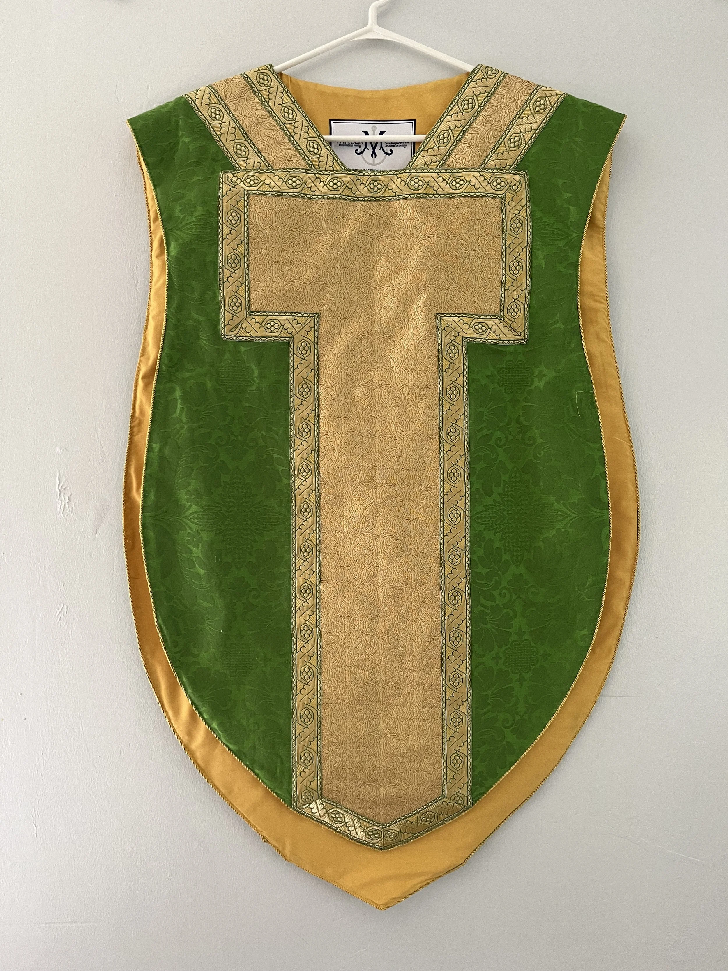A liturgical vestment hanging on a wall, featuring green and gold fabric with intricate embroidery and a shield-shaped design.