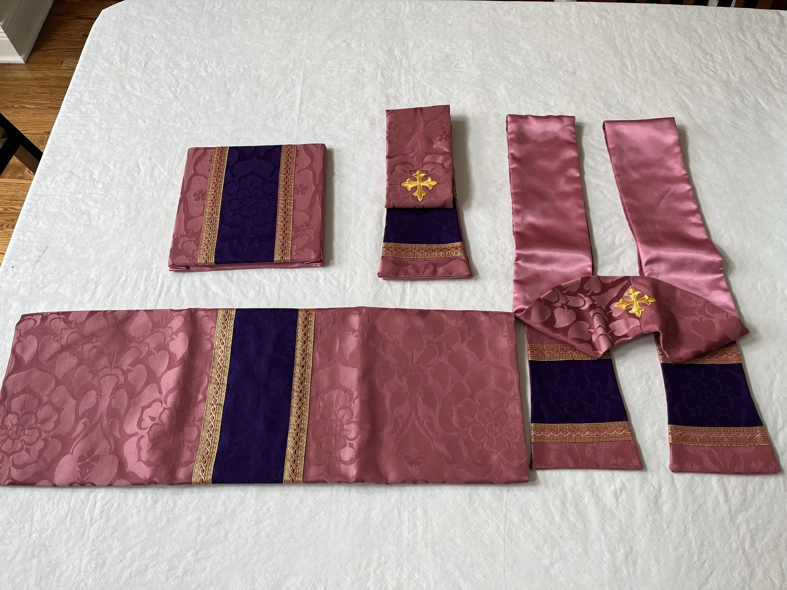 Four pieces of fabric with ornate designs, including one with a gold cross, arranged on a white tablecloth.