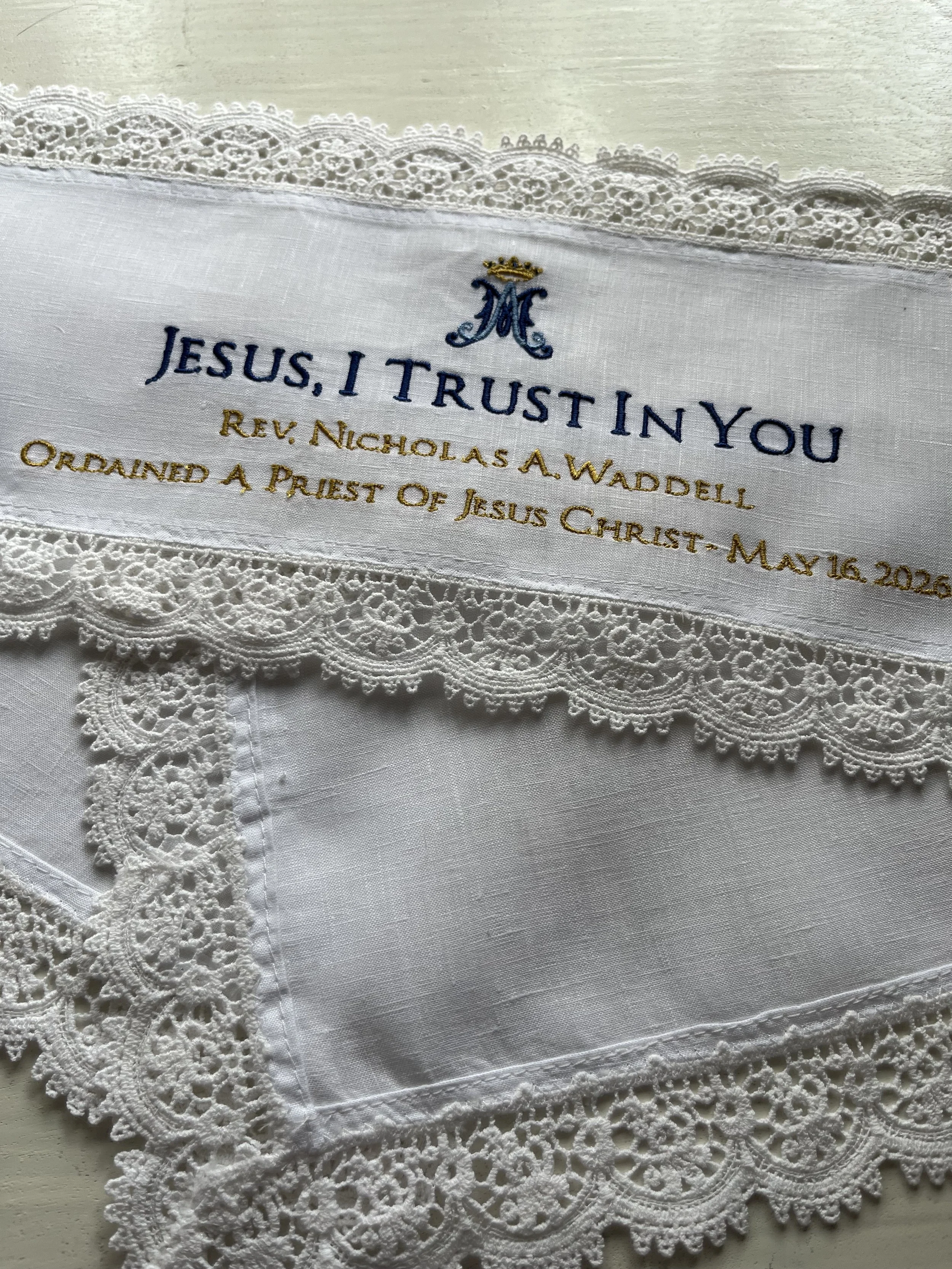 A white cloth with embroidered text reading 'Jesus, I trust in you' and the date 'May 16, 2026', decorative lace trim, and an embroidered monogram with a crown.