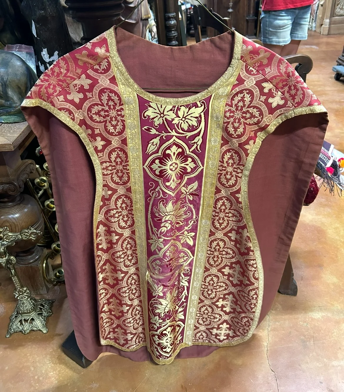 An ornate red and gold vestment with intricate floral embroidery, displayed on a hanger in a store.