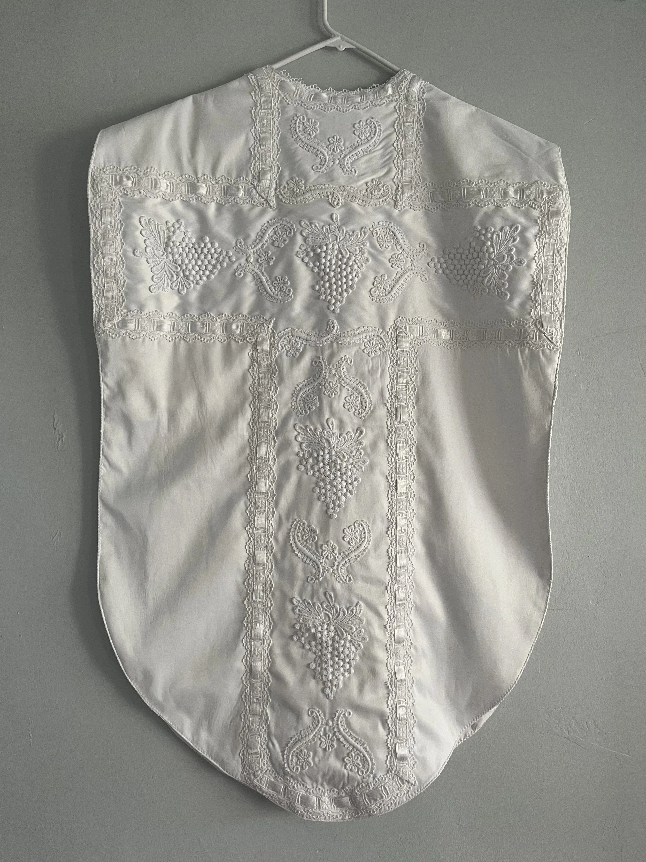 A white embroidered cloth with intricate floral and grapevine patterns hanging on a white hanger against a gray wall.