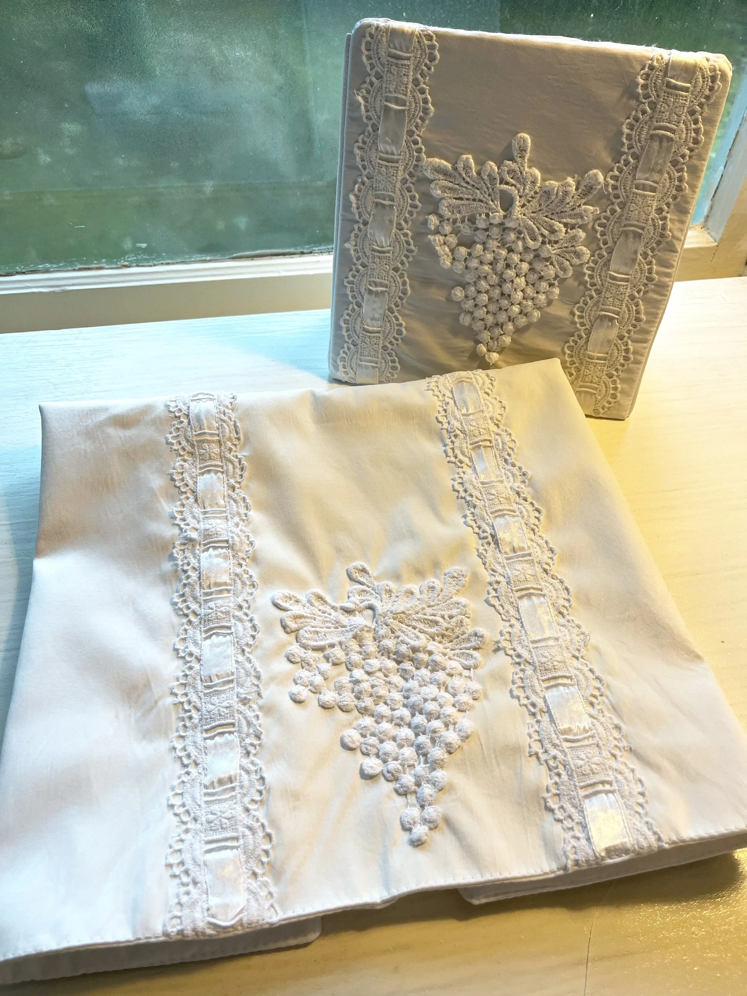 Close-up of a white fabric tablecloth with intricate embroidery and lace details on a wooden surface near a window.
