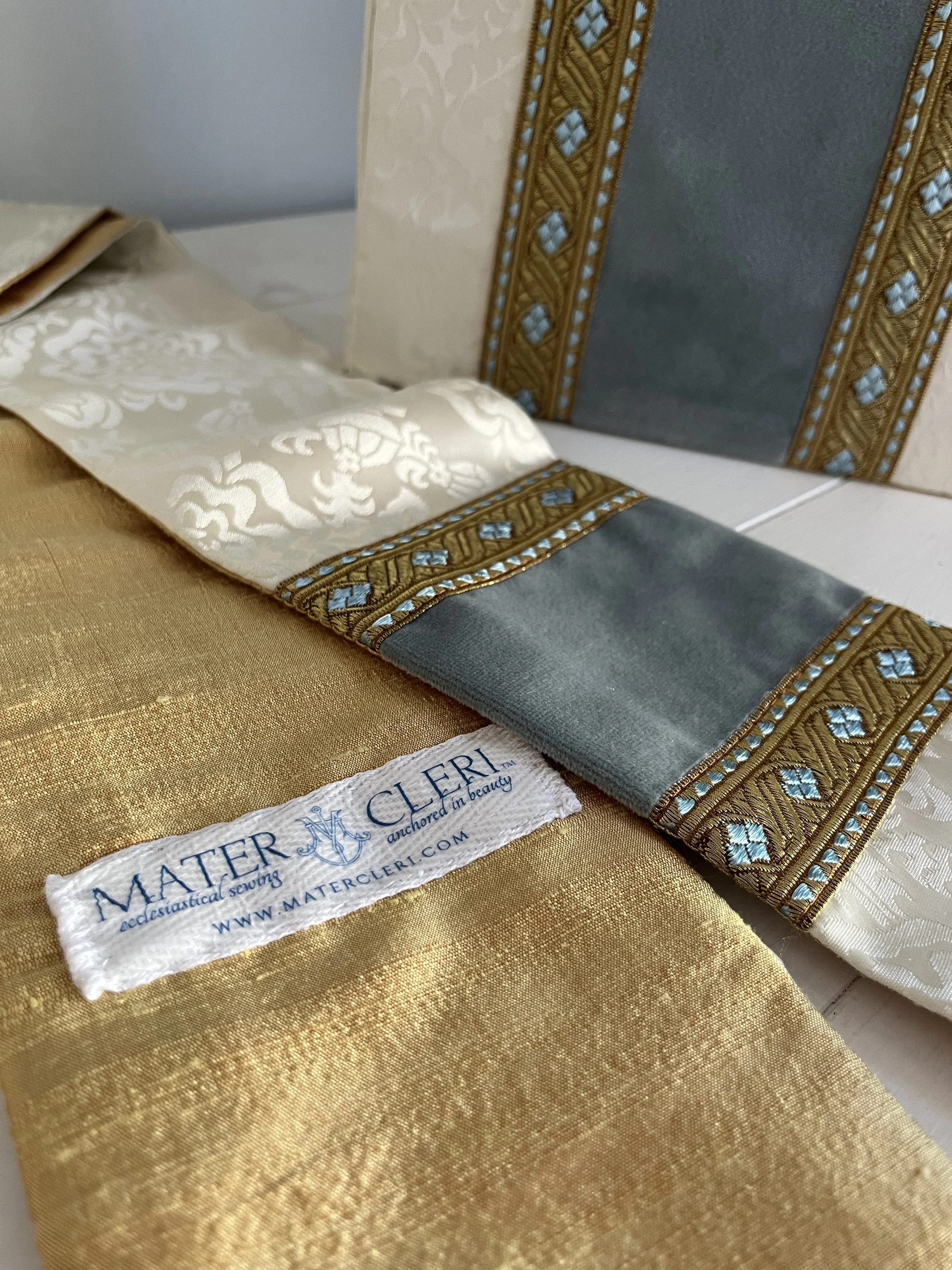 Close-up image of a gold-colored fabric, an embroidered placemat with blue and gold details, a white cloth with intricate embroidery, and a rug cleaning cloth with the logo 'Master M' and the website 'mastermathercli.com'.
