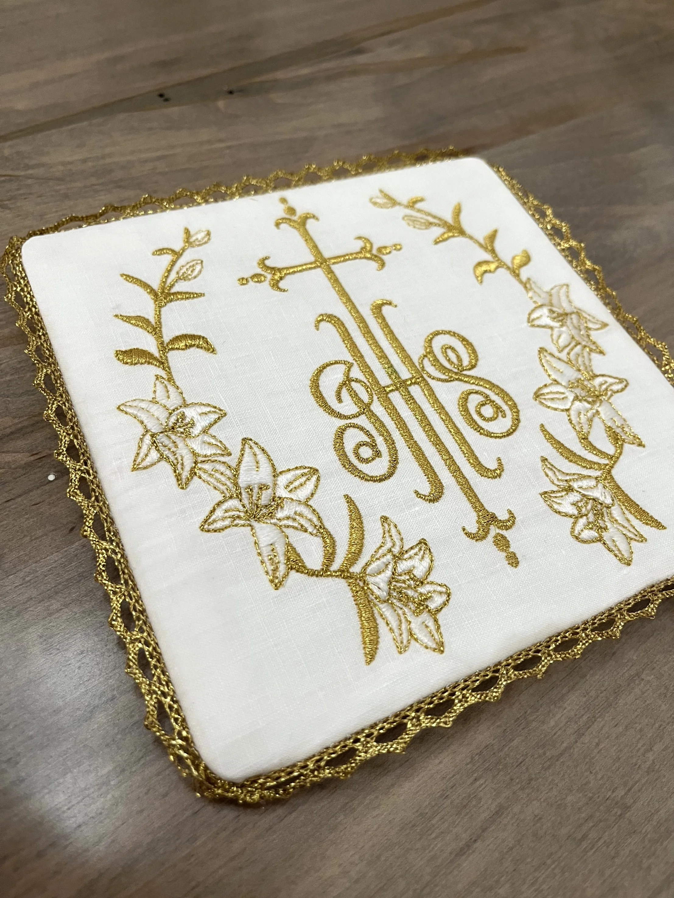 A white cloth embroidered with gold thread featuring floral designs and a central design resembling a musical instrument, bordered with a gold lace trim.