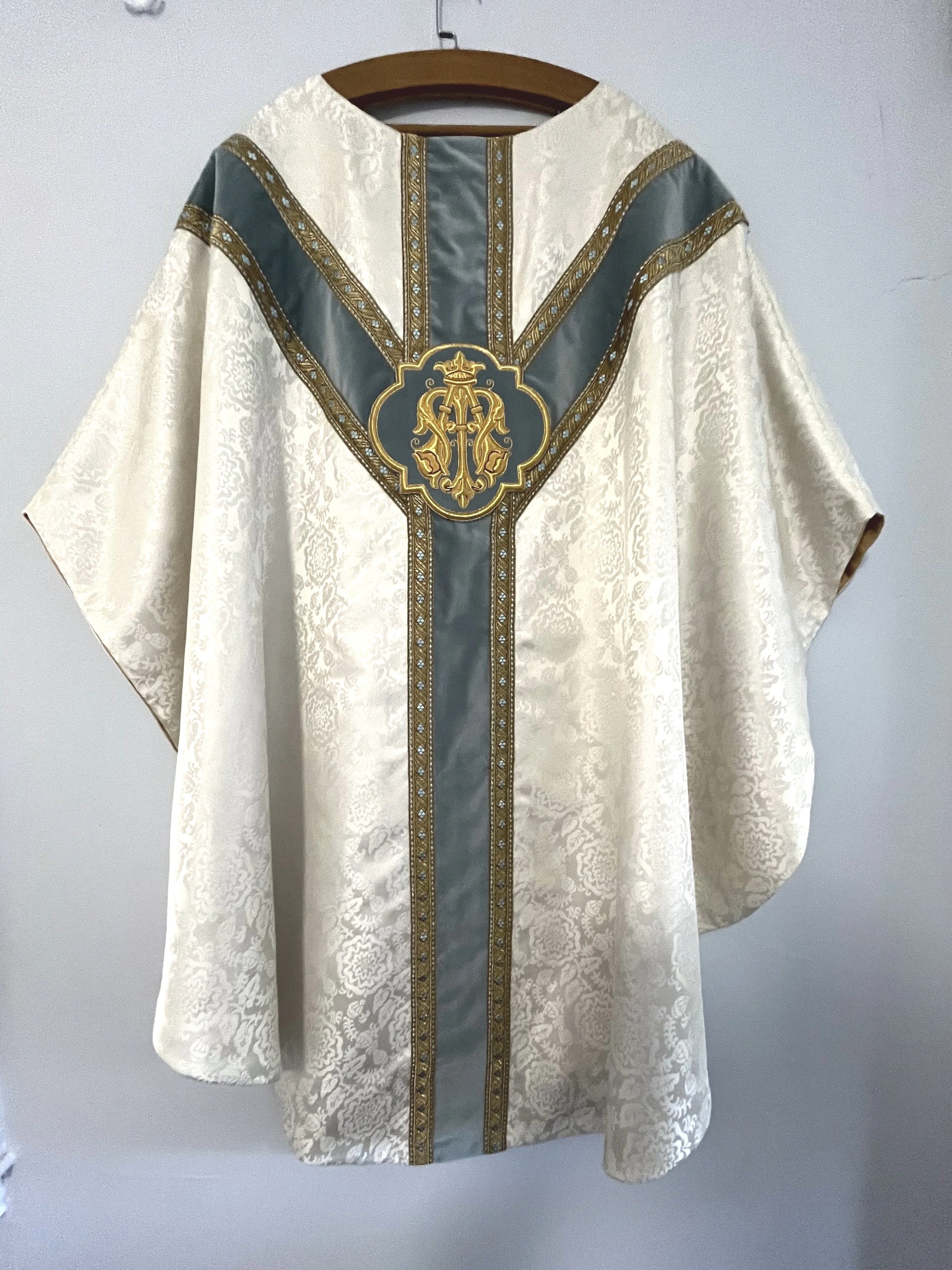 A white religious vestment with intricate gold and silver embroidery, hanging on a wooden hanger against a plain gray wall.