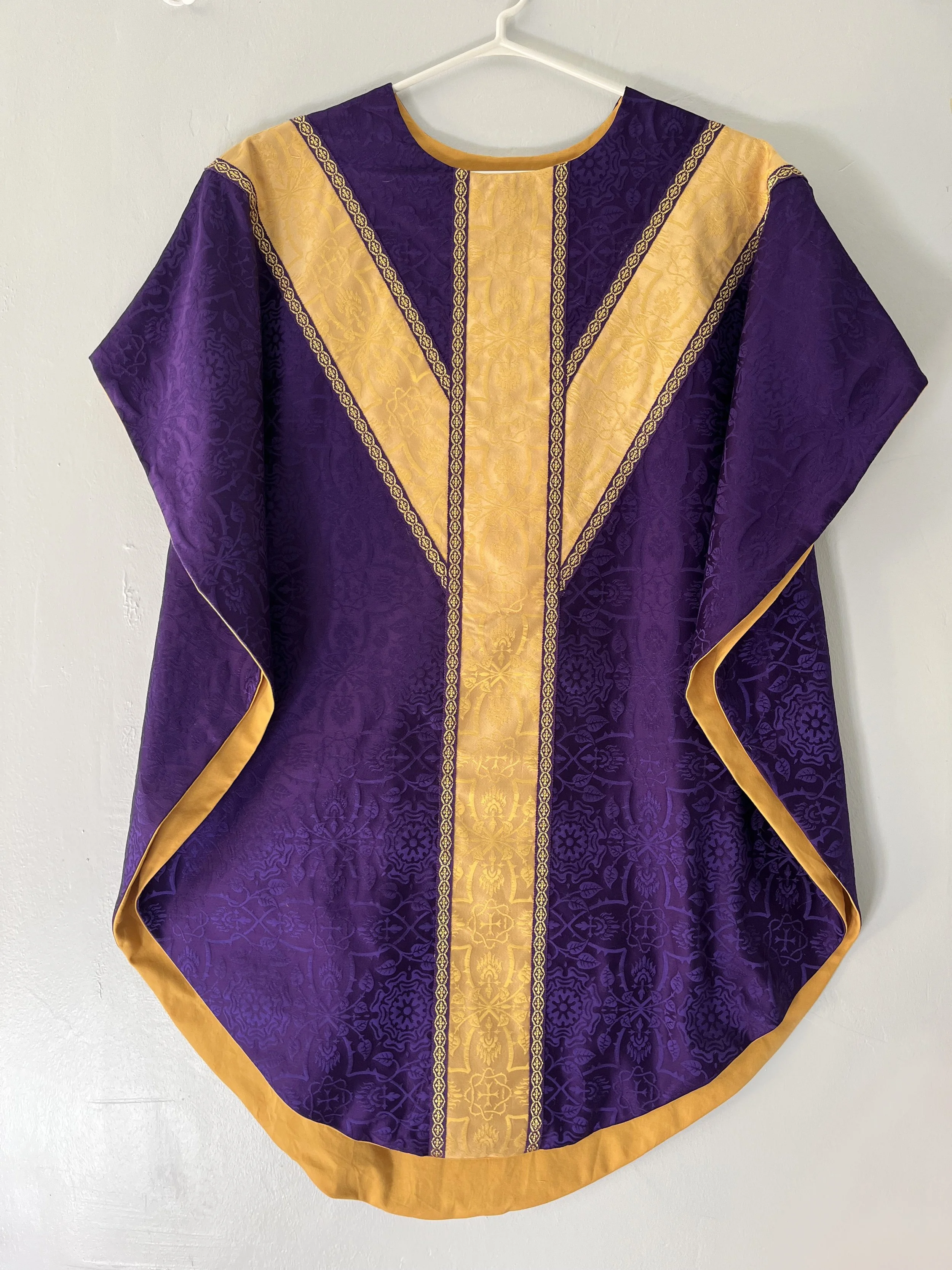 A purple and gold robe with intricate patterns, hanging on a white hanger against a plain white wall.