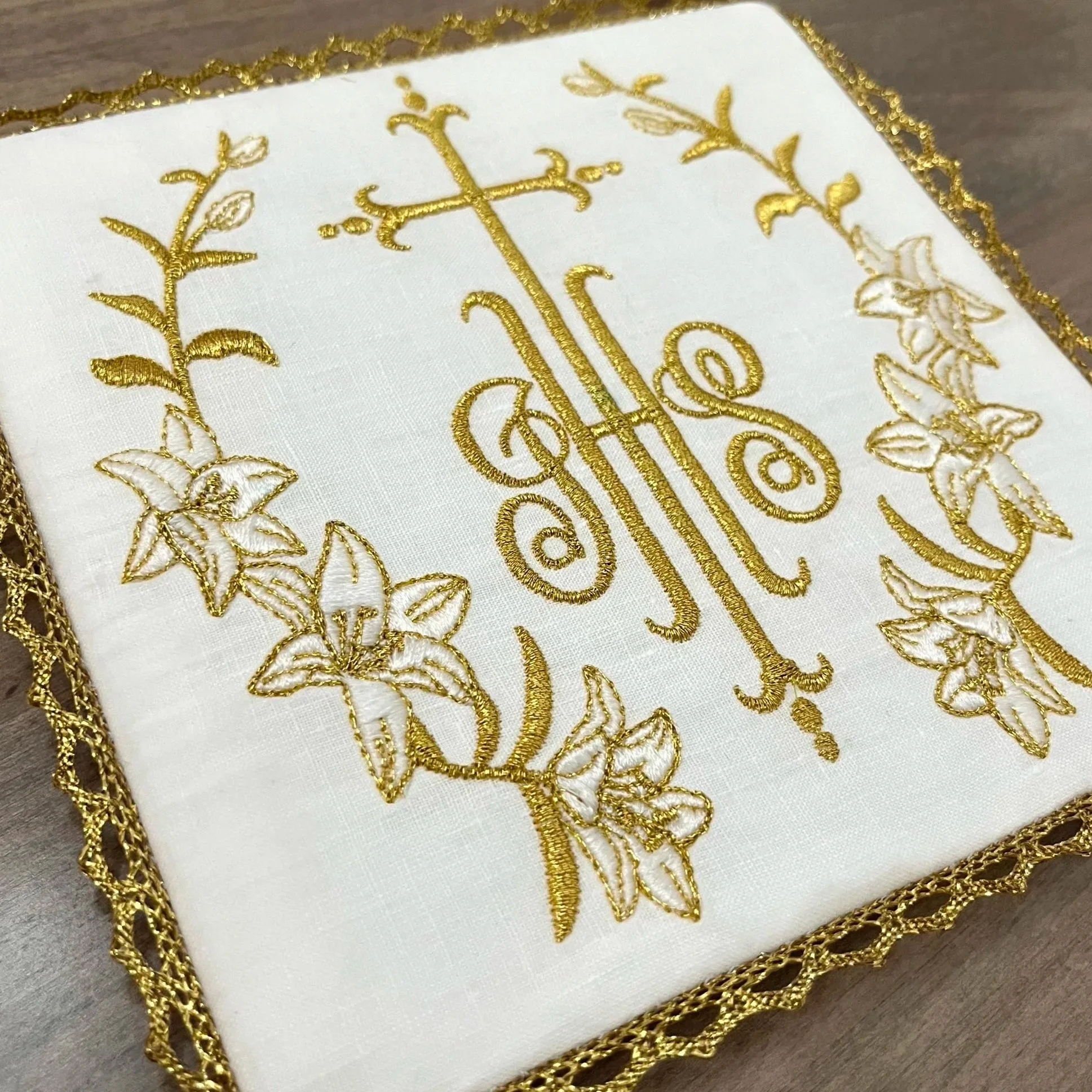 A close-up of white fabric with an ornate, embroidered design featuring gold thread, including floral patterns and a central decorative element, framed by gold braided trim.