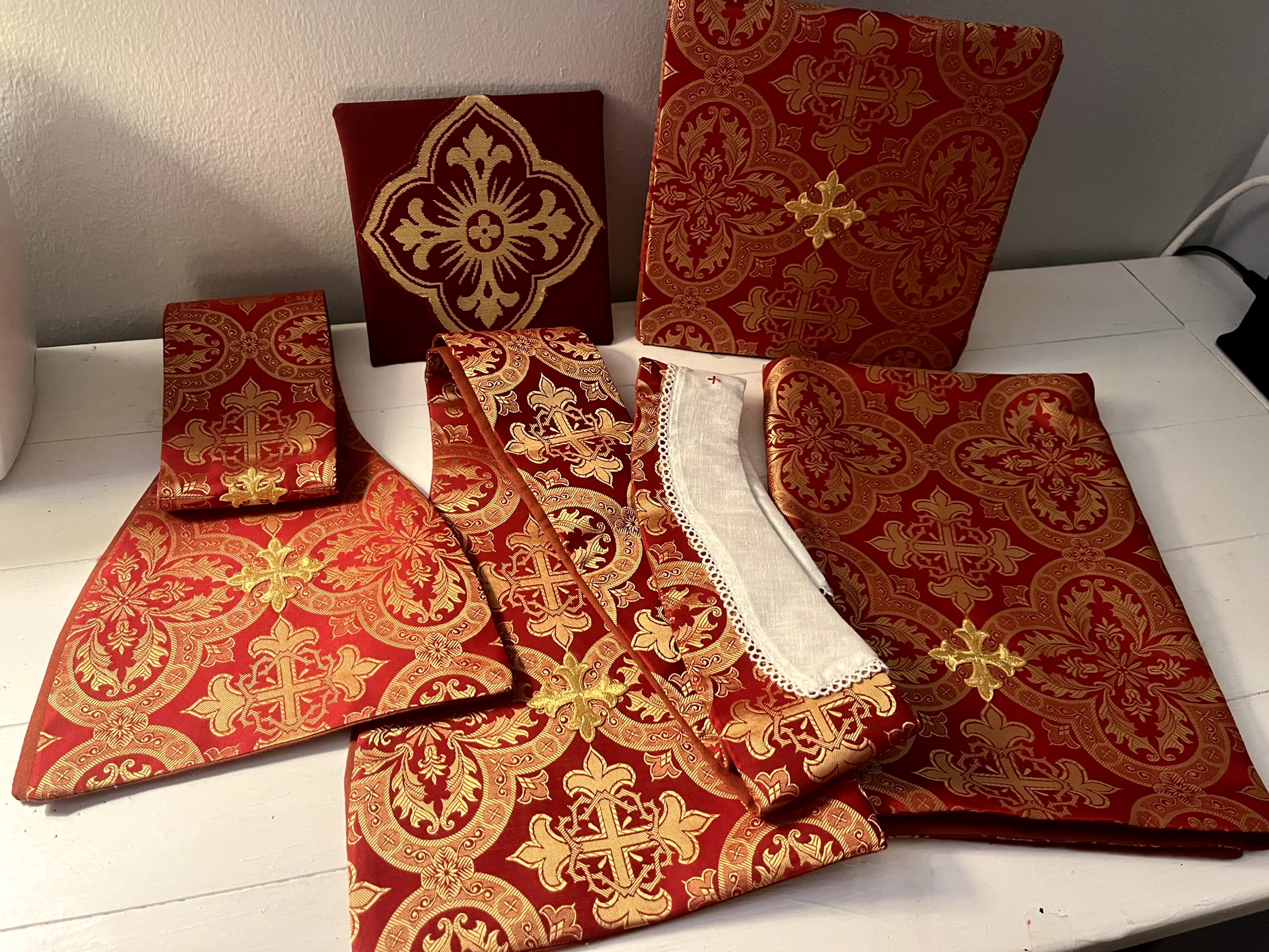 A collection of red and gold religious or ceremonial fabric items on a white surface, including a large folded cloth, a smaller folded cloth, a small bag, and an envelope, all with intricate gold embroidery and patterns.