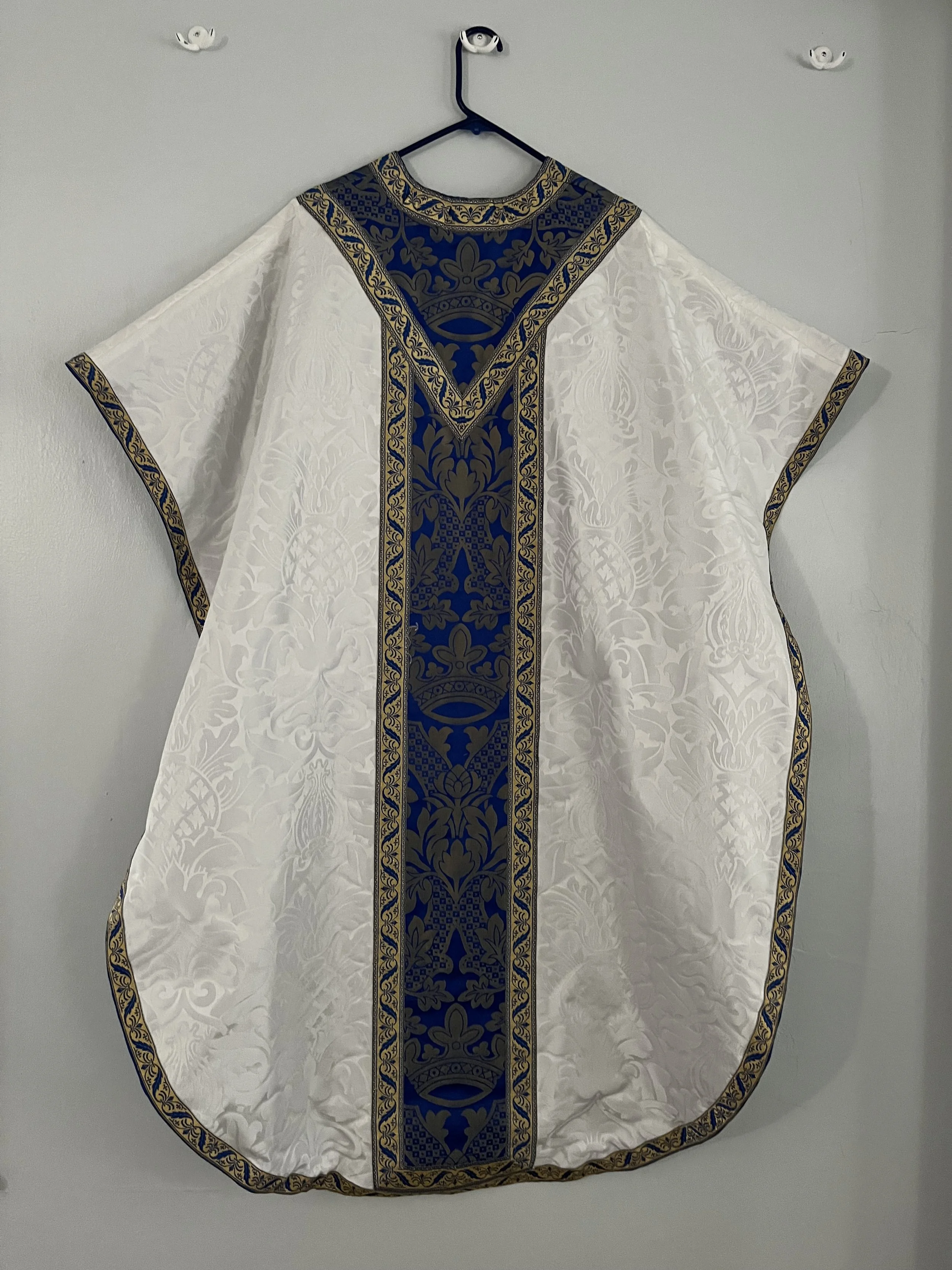 A white clergy robe with ornate gold and blue embroidery hanging on a blue hanger against a plain wall.
