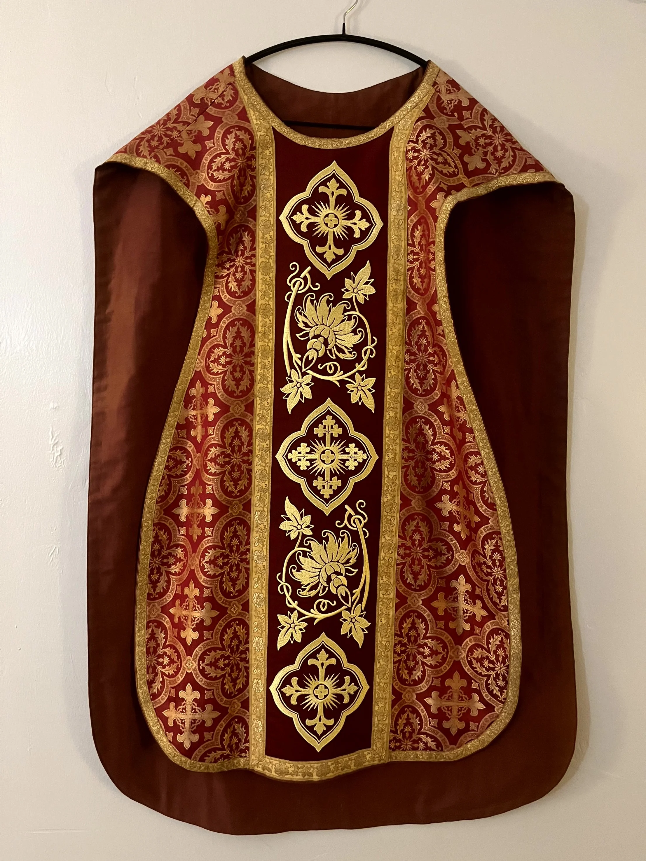 Red and gold ornate religious vestment or stole hanging on a black hanger against a plain wall.