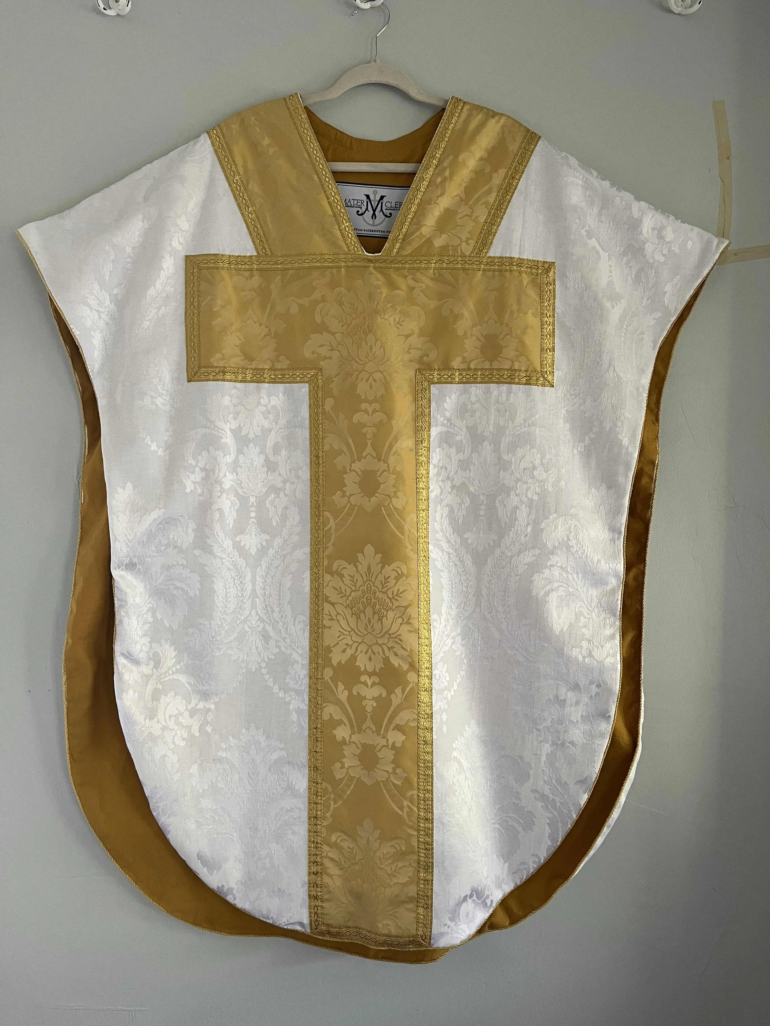 A white and gold liturgical vestment with a large gold cross on the front, hanging on a white hanger against a plain wall.