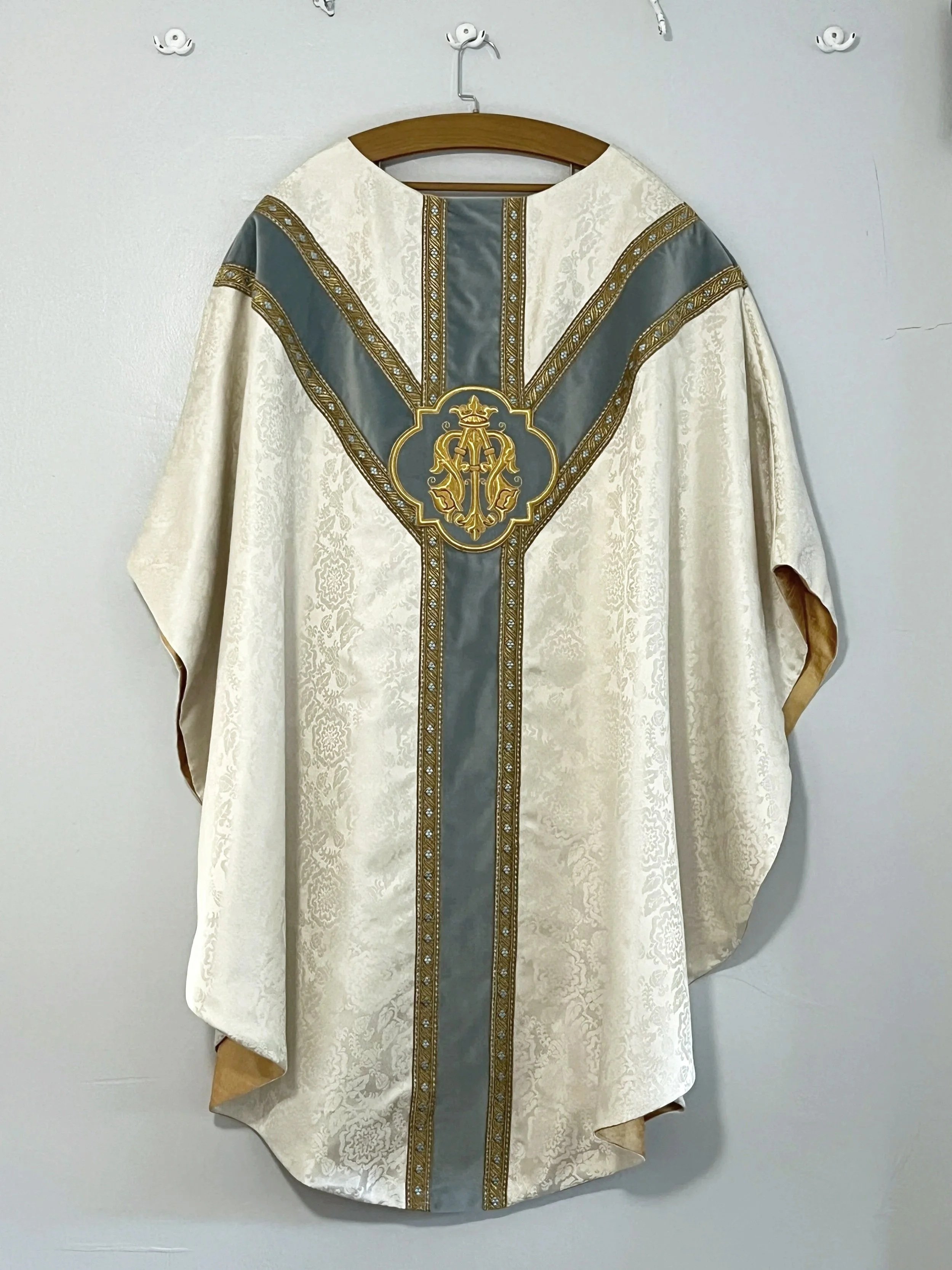 A regal or ceremonial robe with intricate gold and gray designs hanging on a wooden hanger against a plain wall.