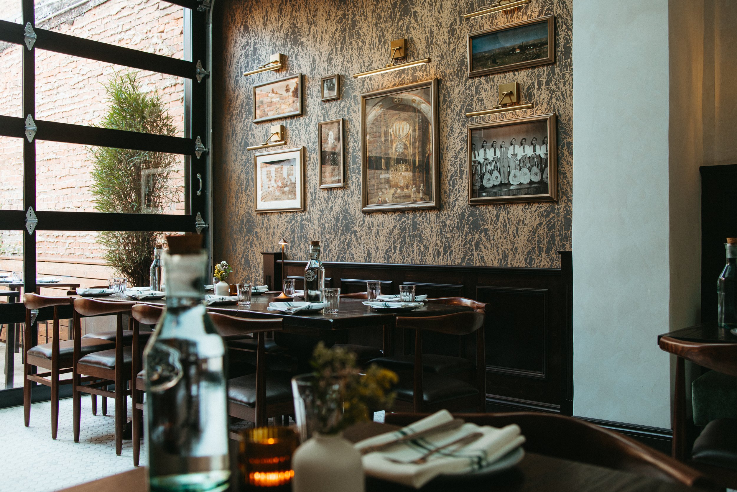 Interior of a restaurant with tables set for dining, large window showing a brick wall and greenery outside, and a wall decorated with framed pictures and art.