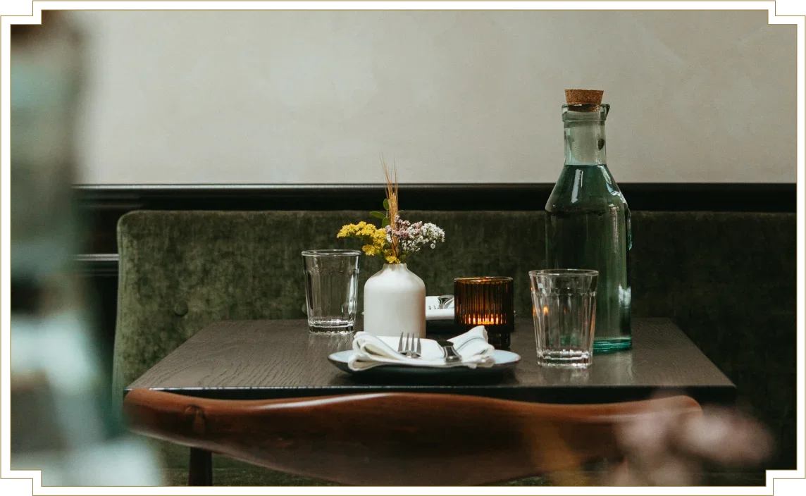 A table setting with a small vase of flowers, two glasses, a water bottle with a cork, a candle, and utensils on a plate, on a dark wooden table in a cozy restaurant or cafe.