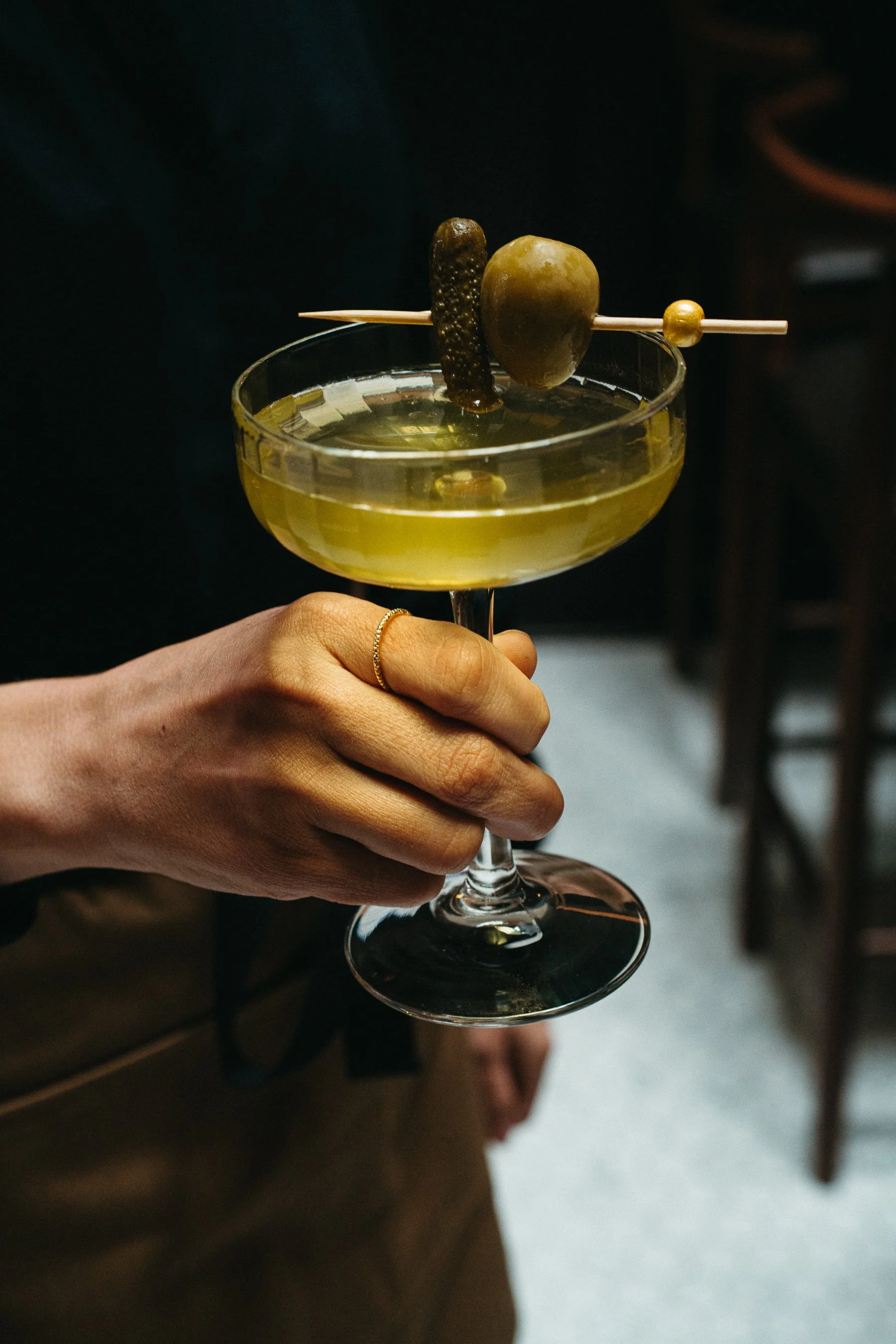 A person holding a cocktail glass with a green olive and a small pickle skewer garnished with a small green olive.