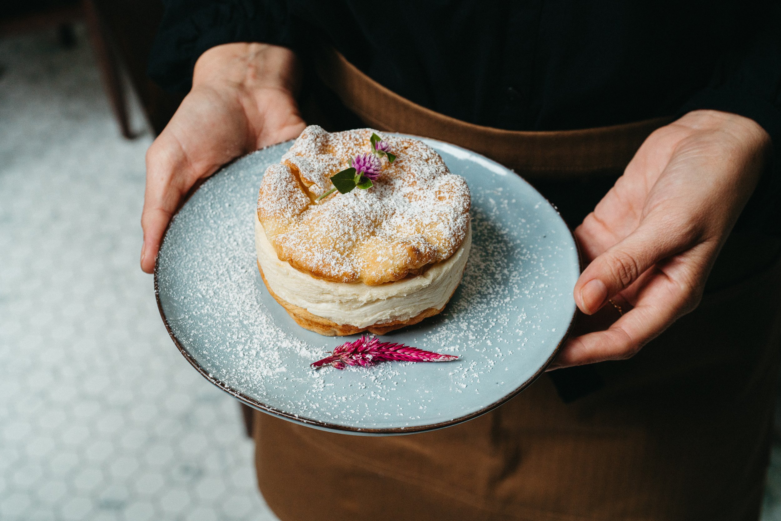 A person holding a cake on a blue plate. The cake is topped with powdered sugar, a small pink flower, green leaves, and a pink flower sprig.