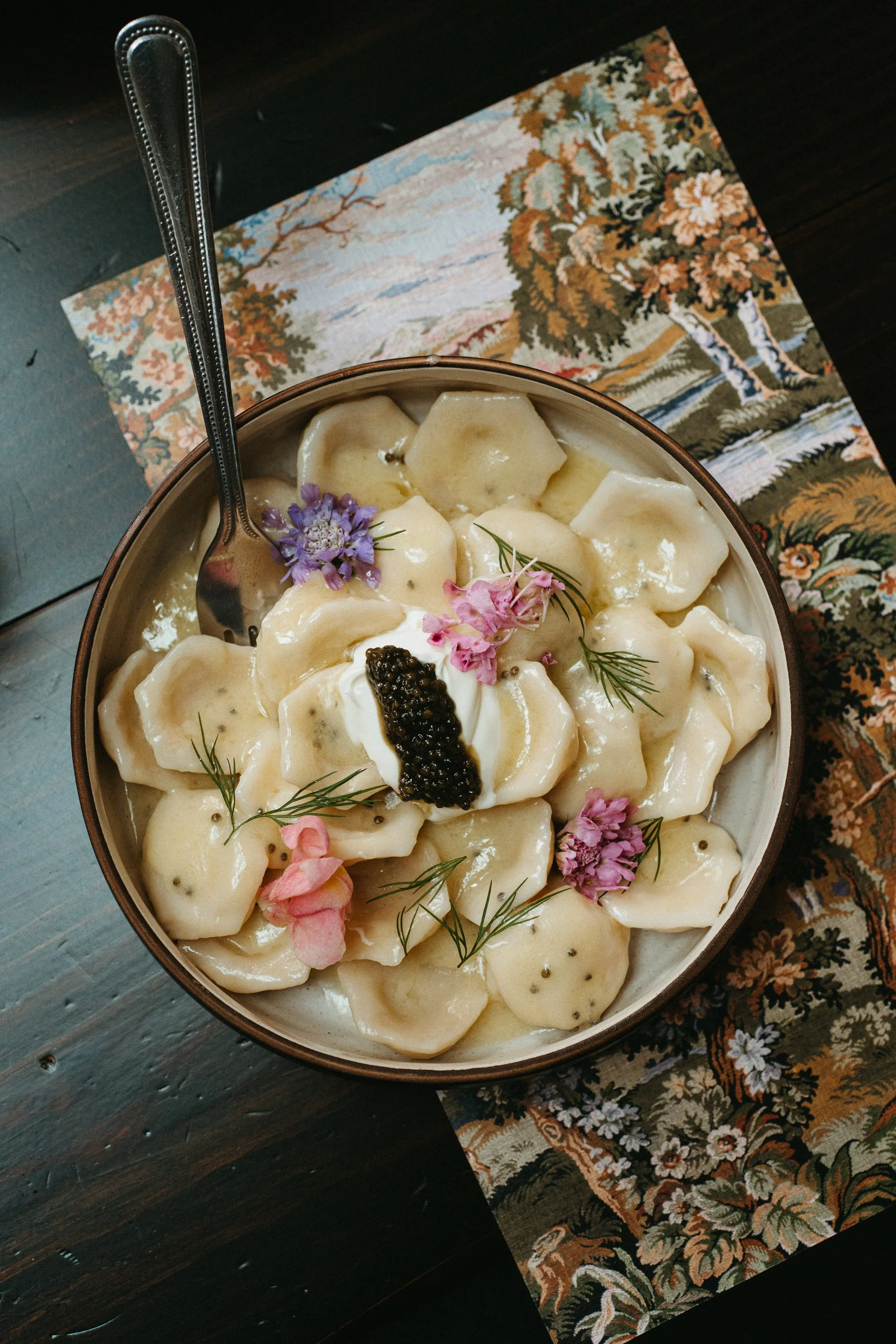 A bowl of potato gnocchi topped with black caviar, herbs, and edible flowers, placed on a floral table runner on a dark wooden table.