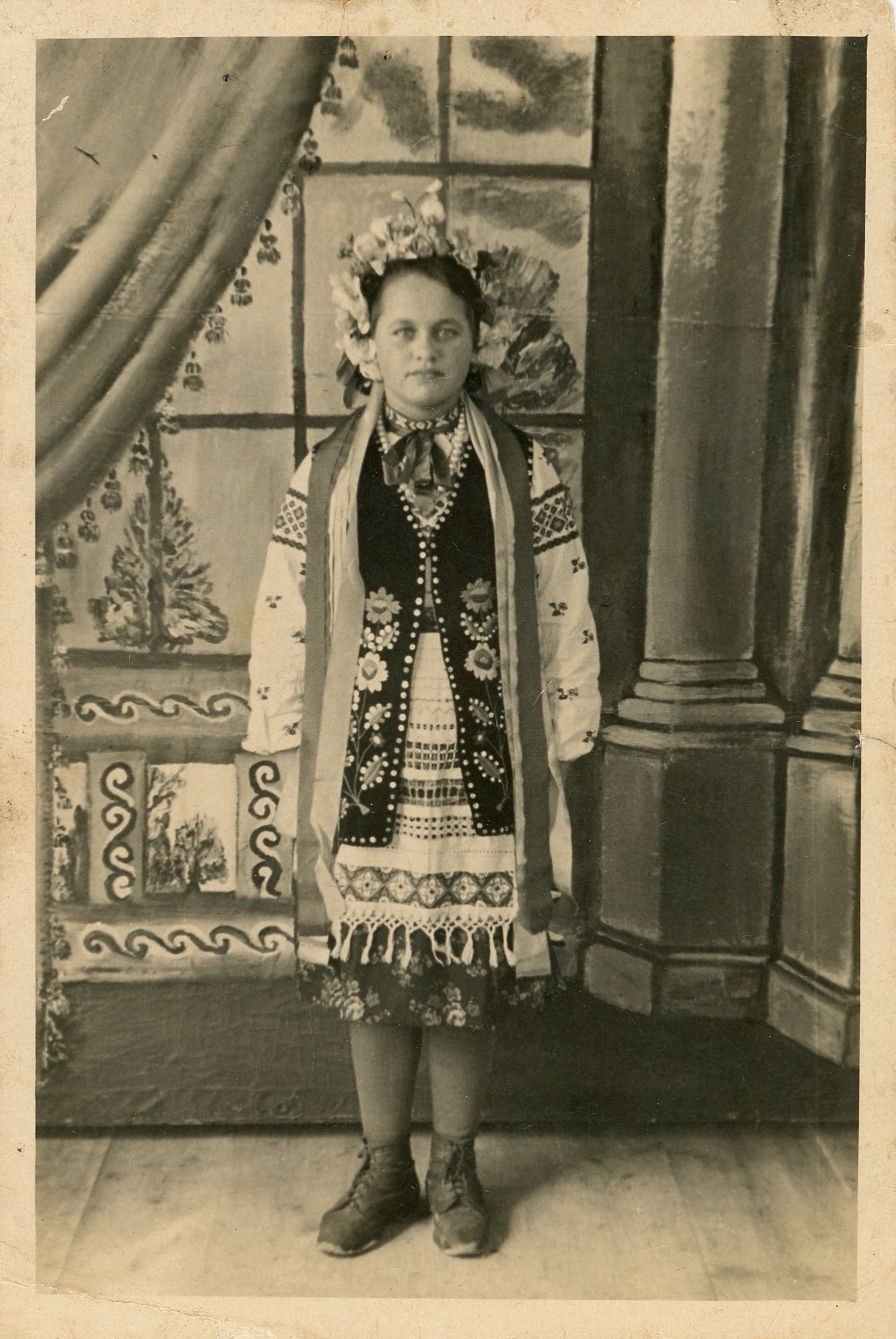 A woman dressed in traditional folk costume, standing indoors in front of a decorated backdrop with paintings and architectural details.
