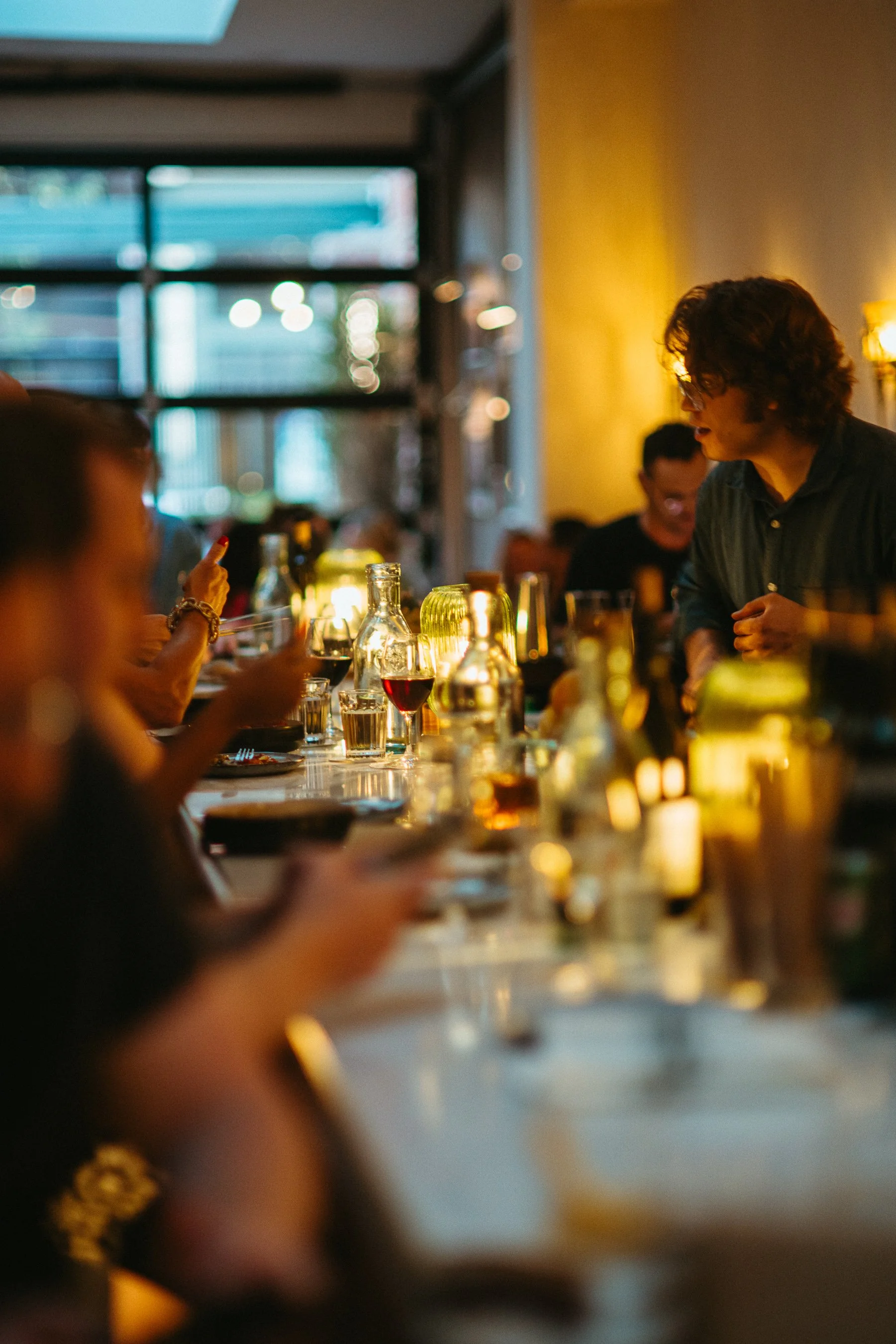 People socializing and enjoying drinks in a dimly-lit restaurant or bar with warm lighting and large windows.