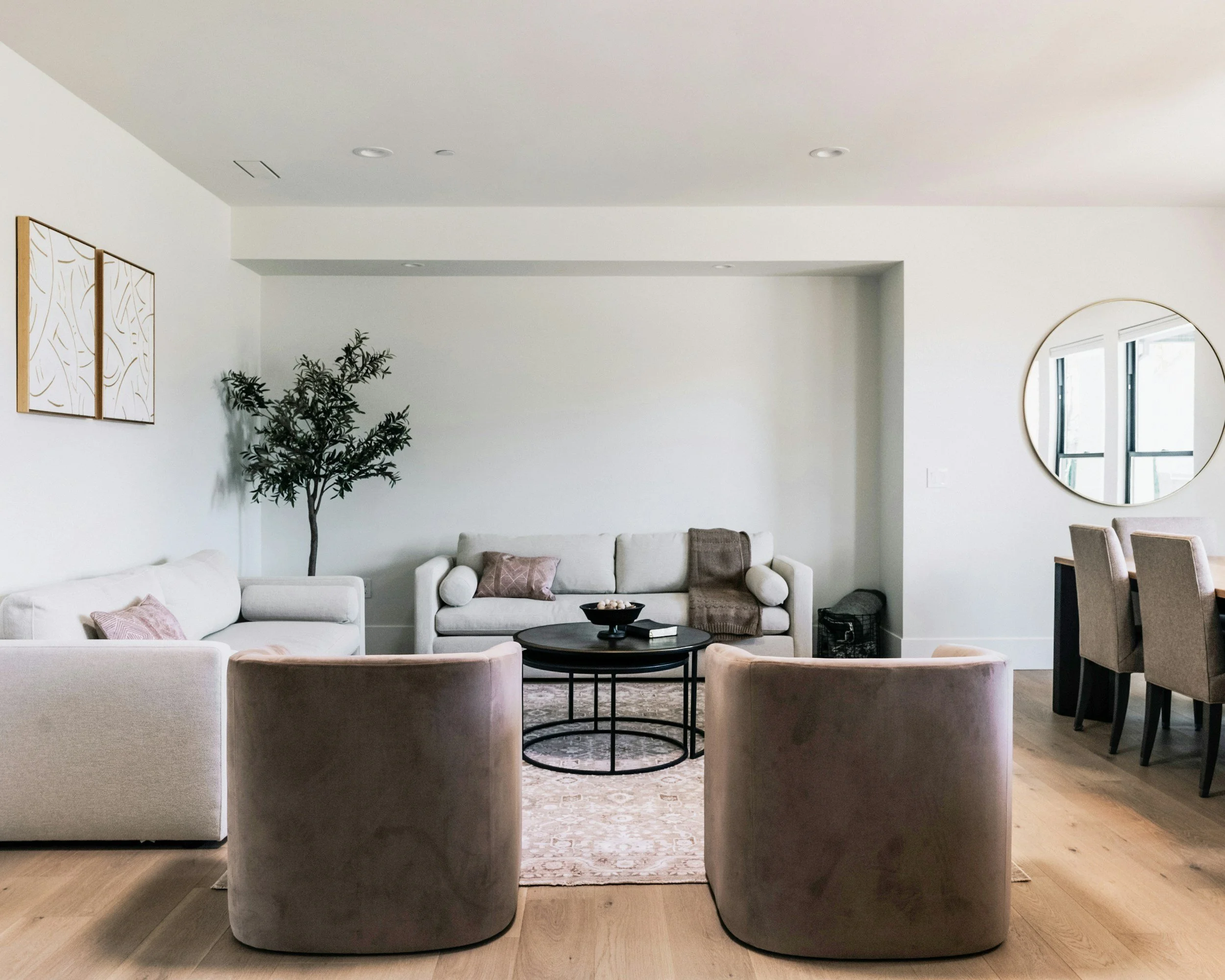 Modern living room with white sofas, pink pillows, wooden side tables, and a round mirror on the wall.