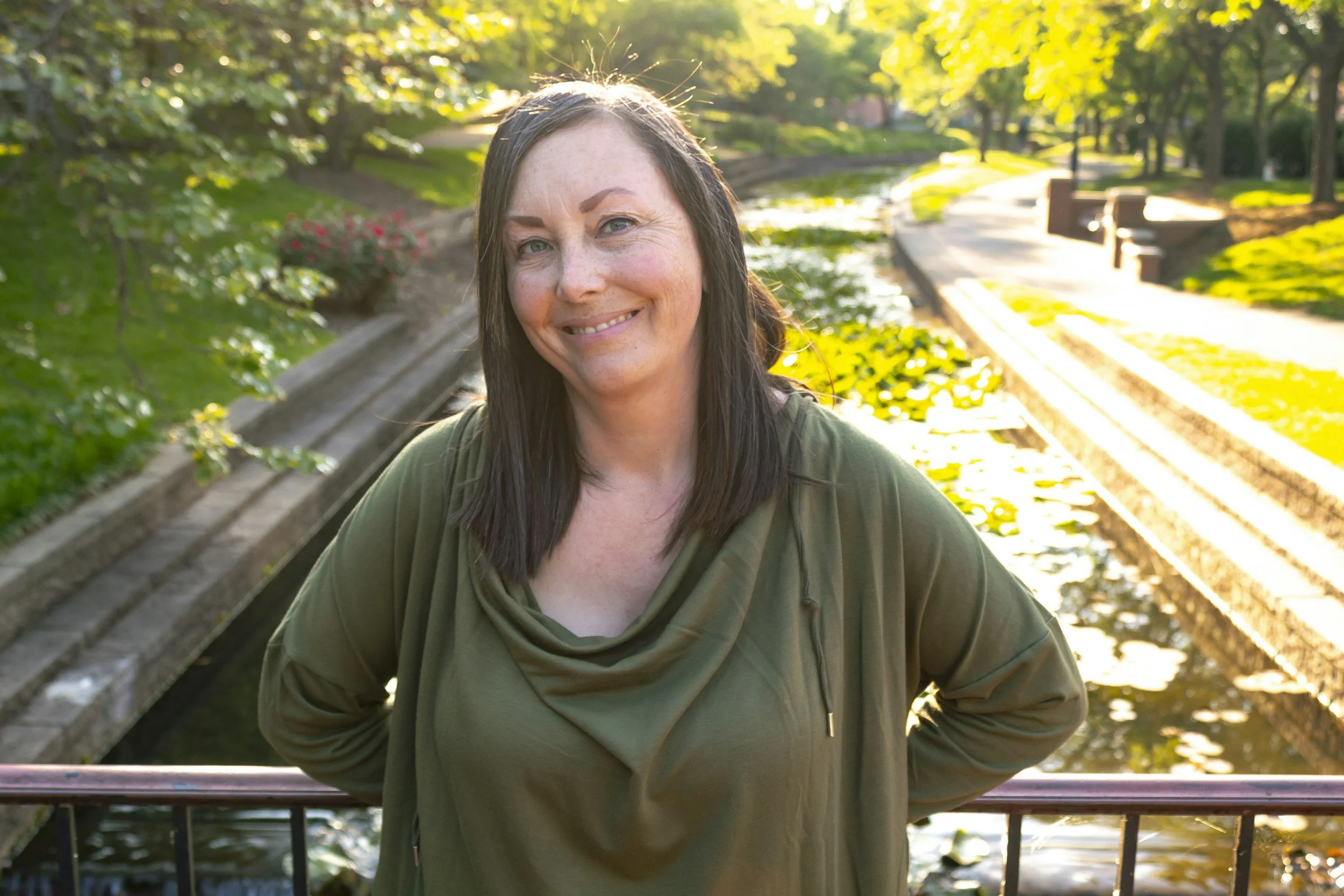 A woman with long dark hair and a warm smile standing outdoors on a bridge in a park during sunset, with green trees and a small waterway in the background.
